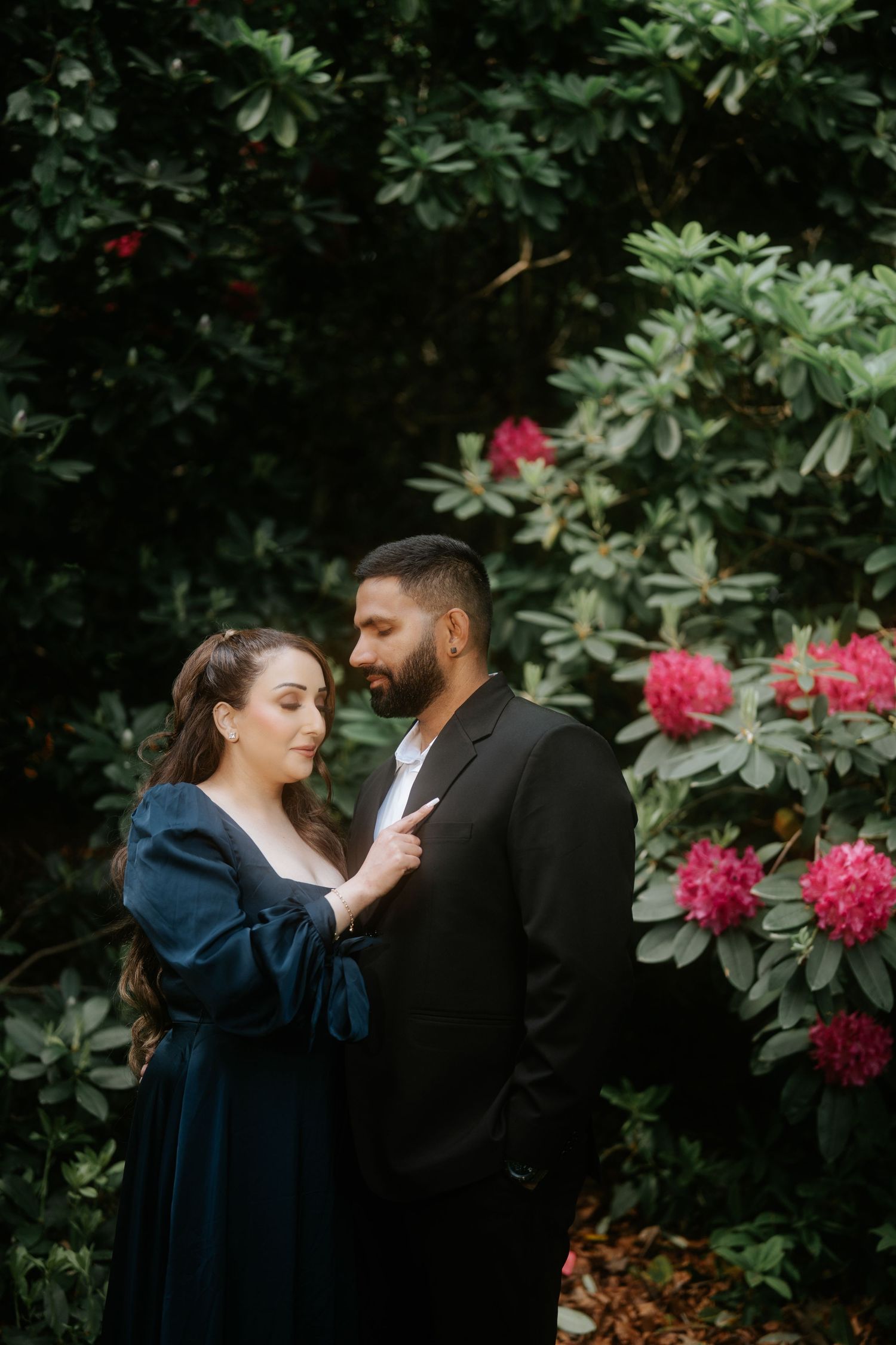 Couple in elegant attire embracing among lush green bushes and pink rhododendron flowers in a garden.