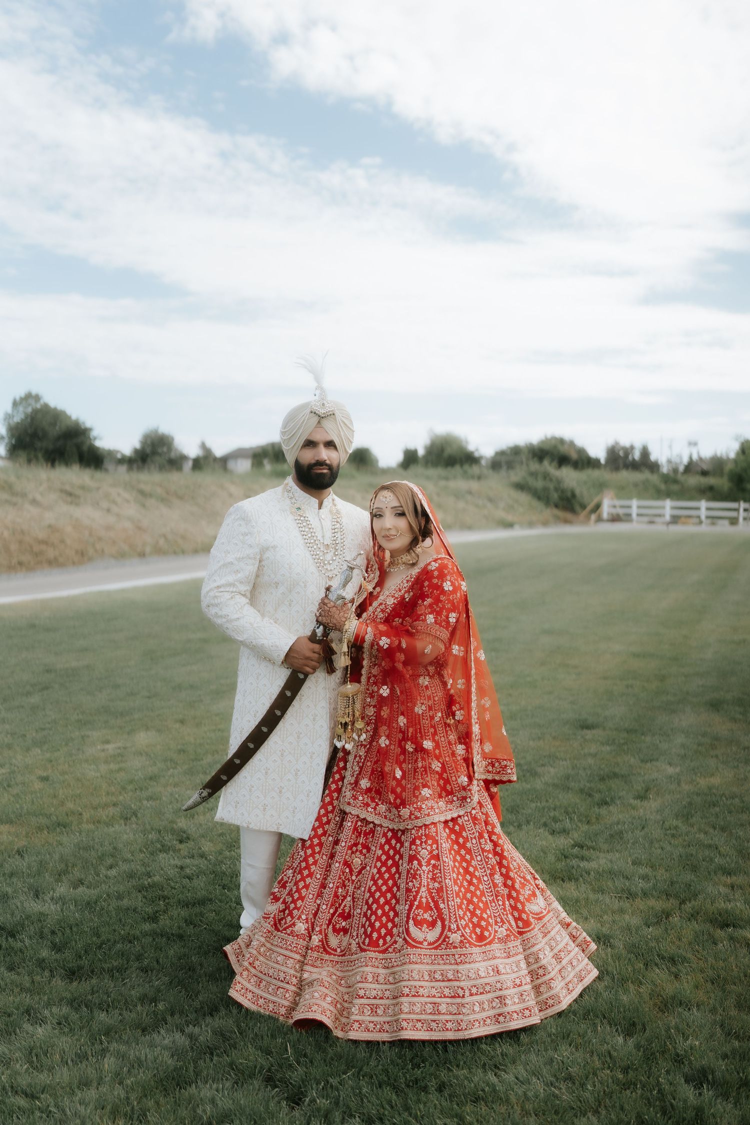 Sikh bride in red lehenga and groom in white sherwani and turban pose together on green lawn at outdoor wedding.