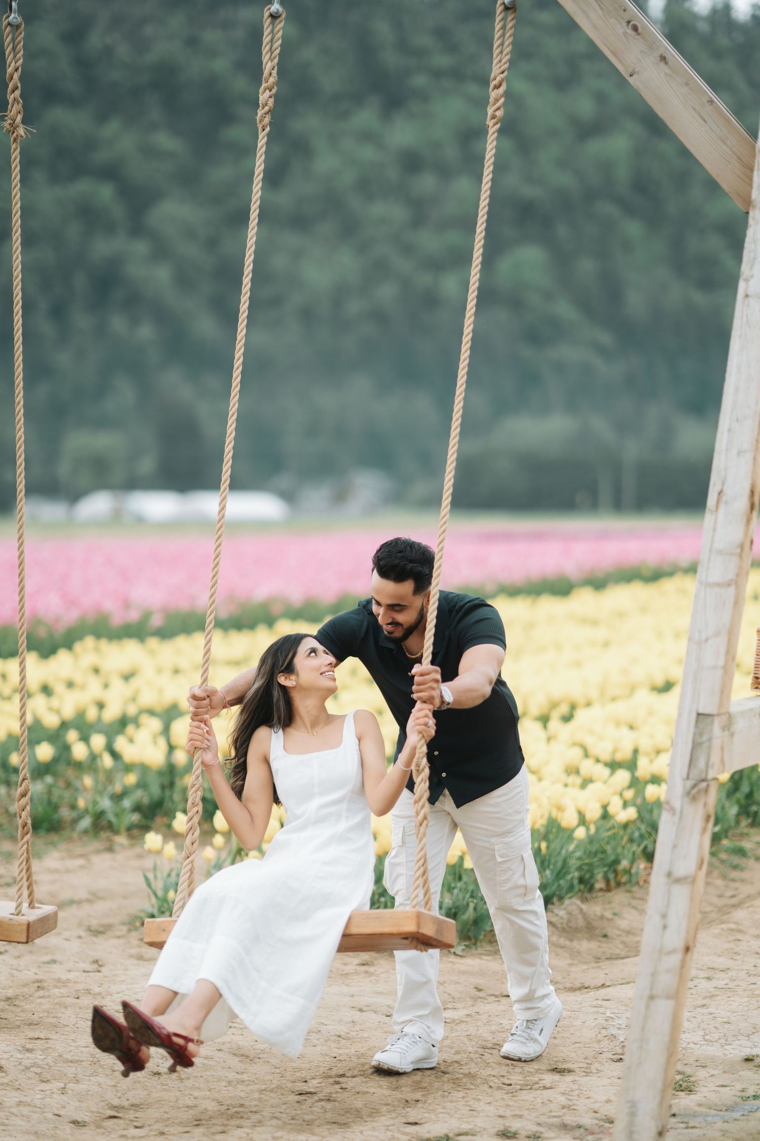 Couple playfully swinging on wooden swings in a tulip field with yellow and pink flowers in the background.
