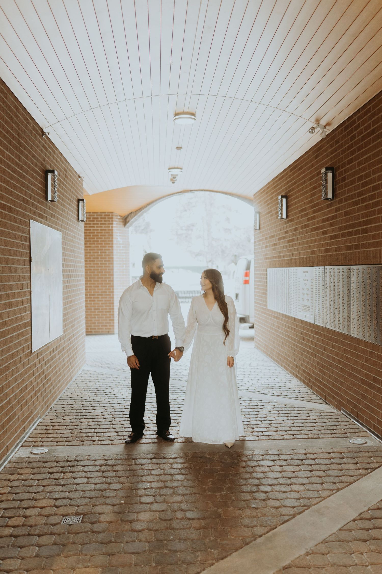A couple walks together in a long brick hallway with white ceiling panels.