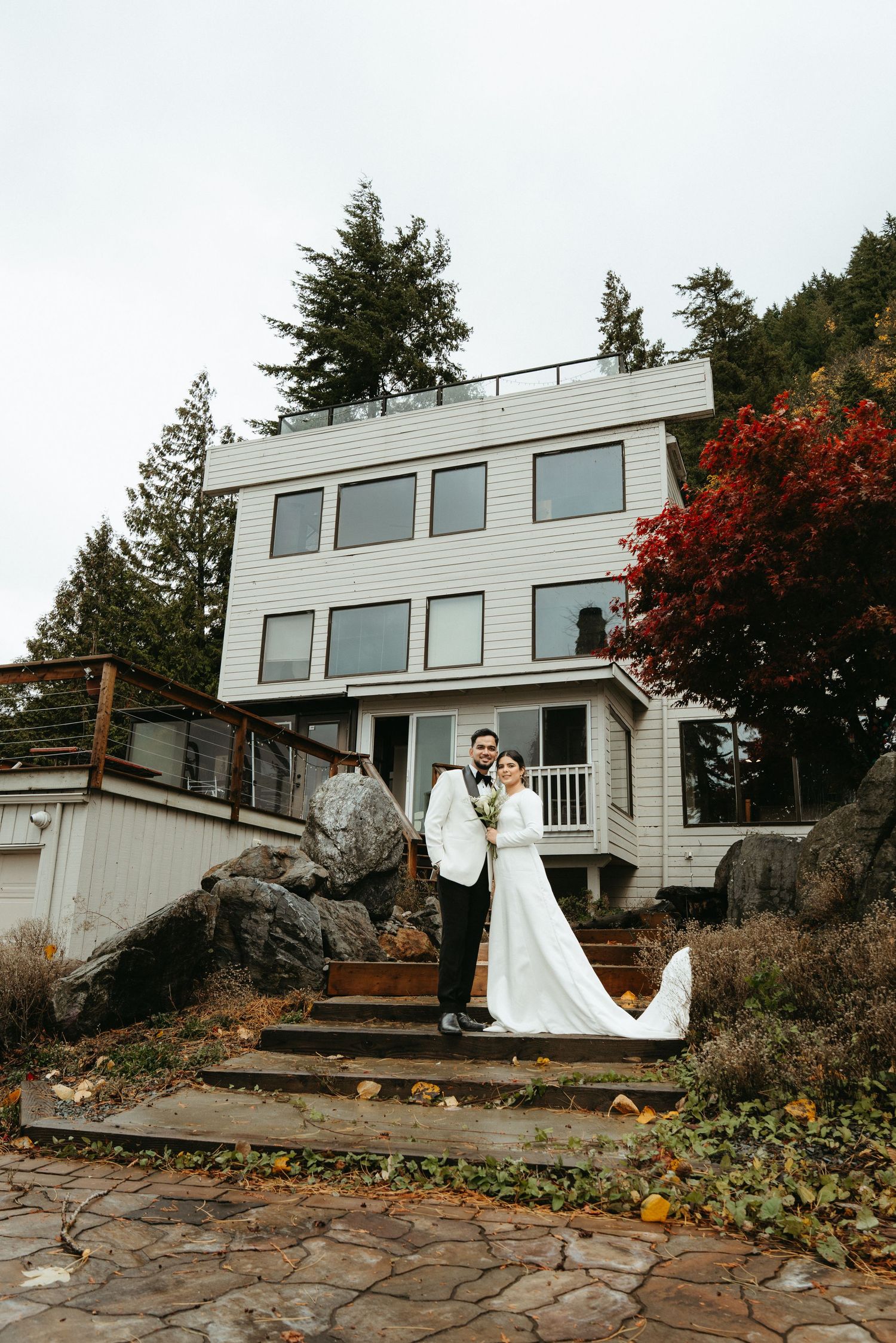 A modern coastal house with large windows sits atop rocky terrain as a couple celebrates their wedding day outdoors.