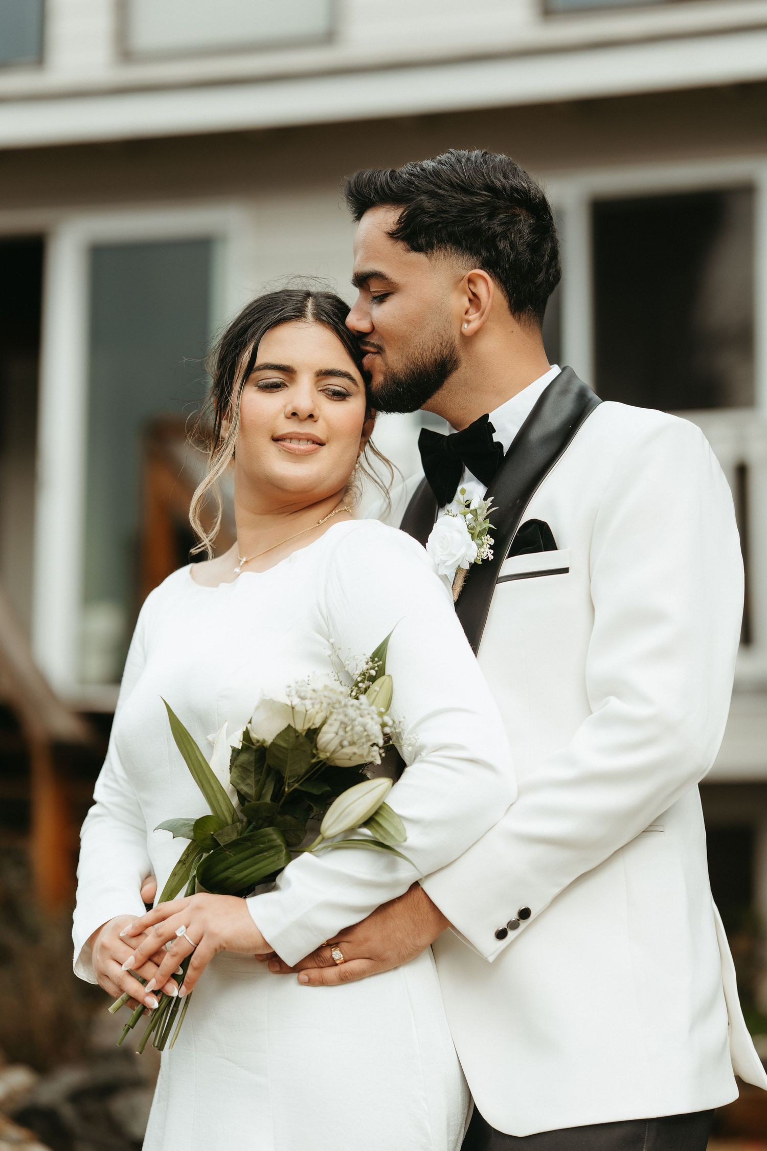 A couple in wedding attire share an intimate moment, with the bride in white dress holding a bouquet and groom in white tuxedo.