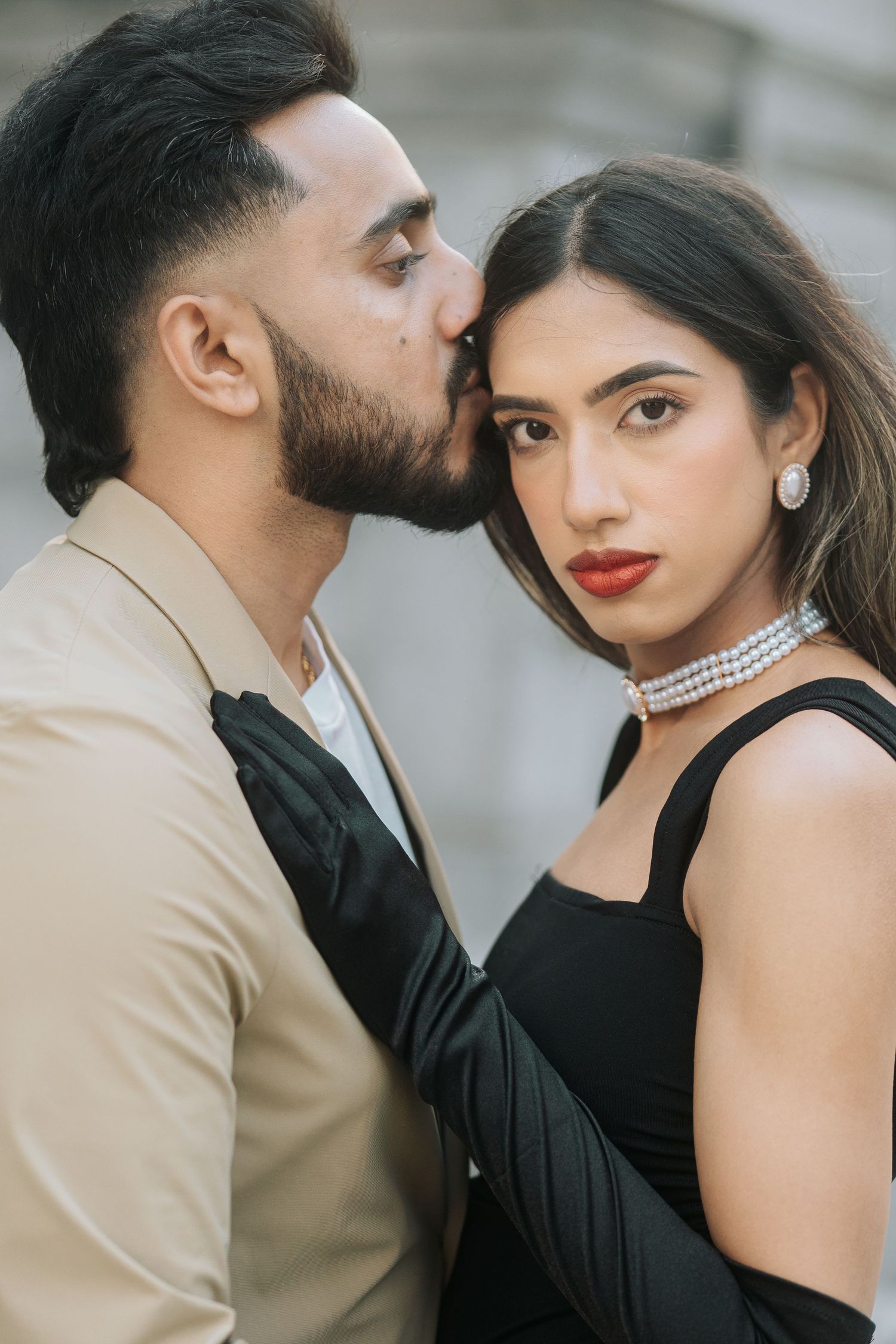 A romantic couple in elegant attire share an intimate moment, with her in black and him in beige.