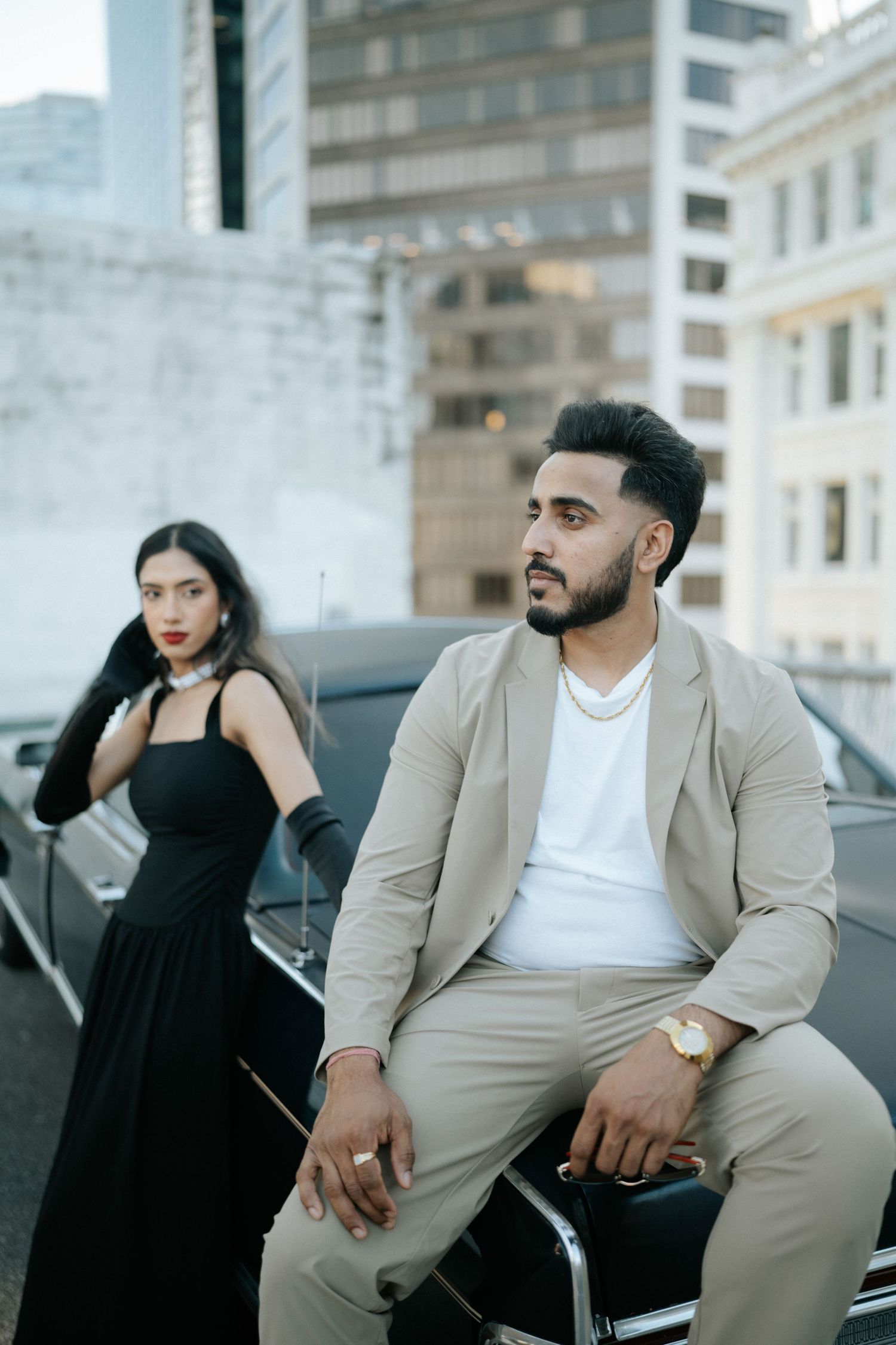 Stylish couple in formal attire pose against urban backdrop with modern buildings and architecture.