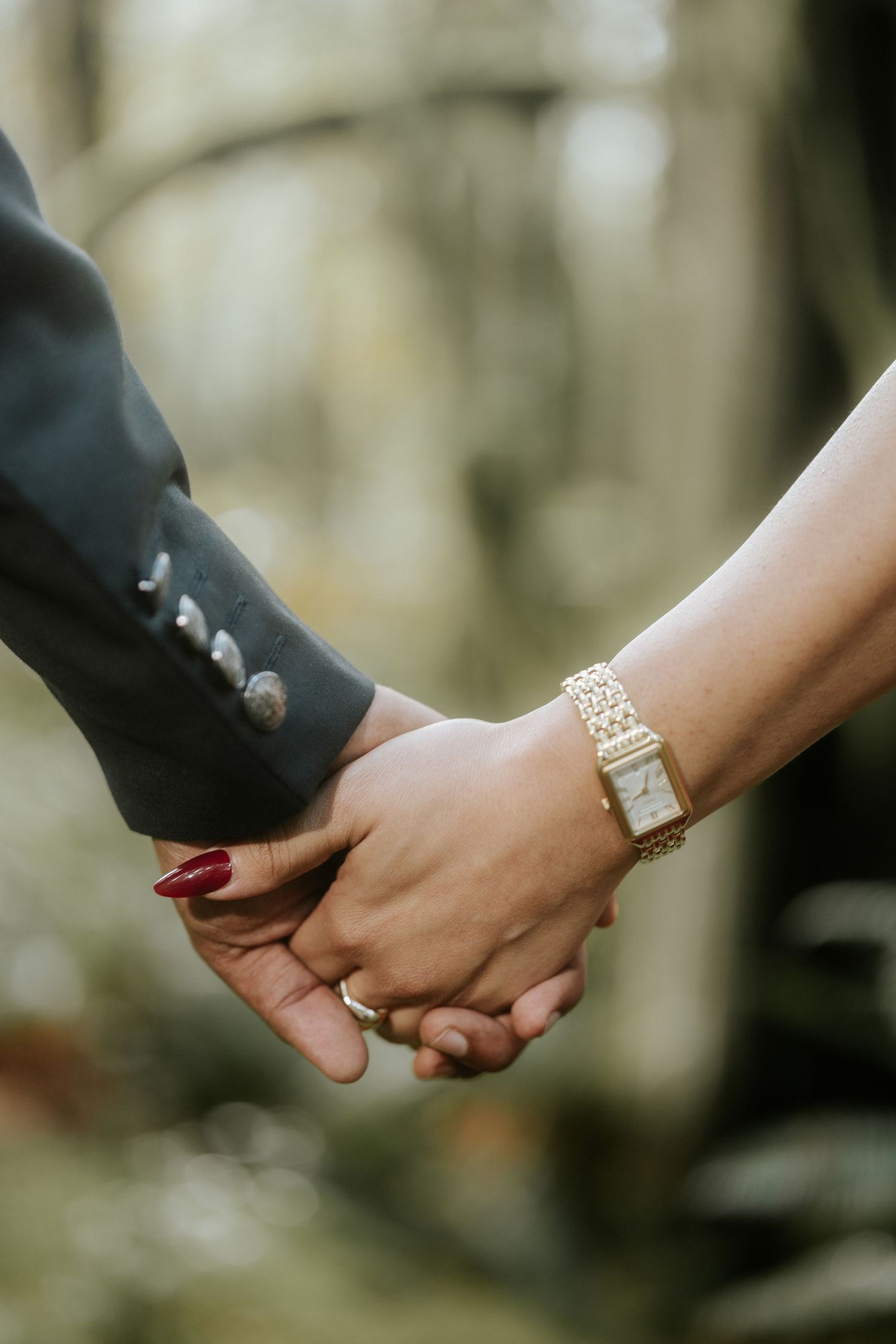 Close-up of joined hands showing jewelry and red nail polish in soft forest light.