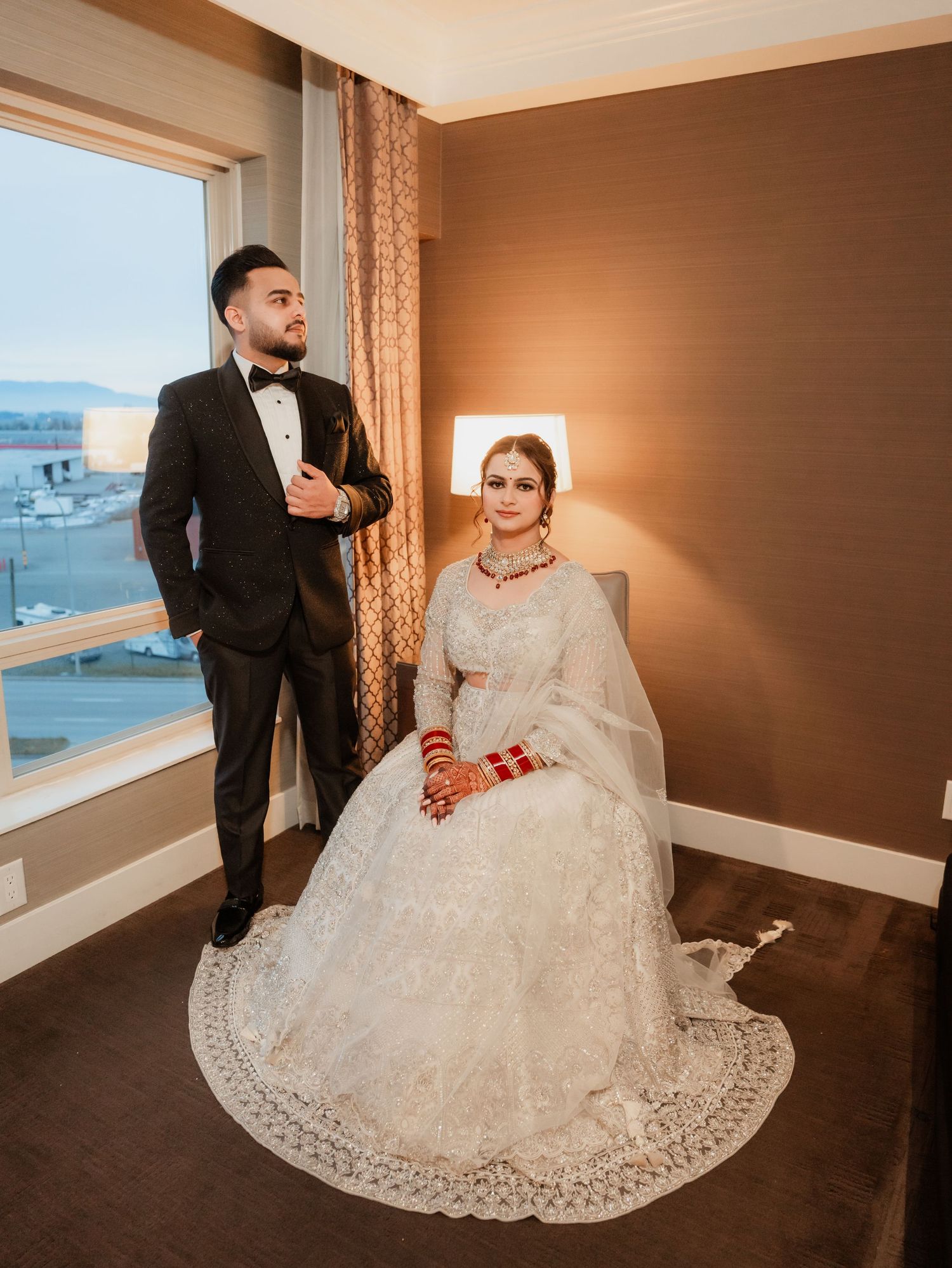 A couple in formal attire poses in hotel room with scenic window view and warm lighting.