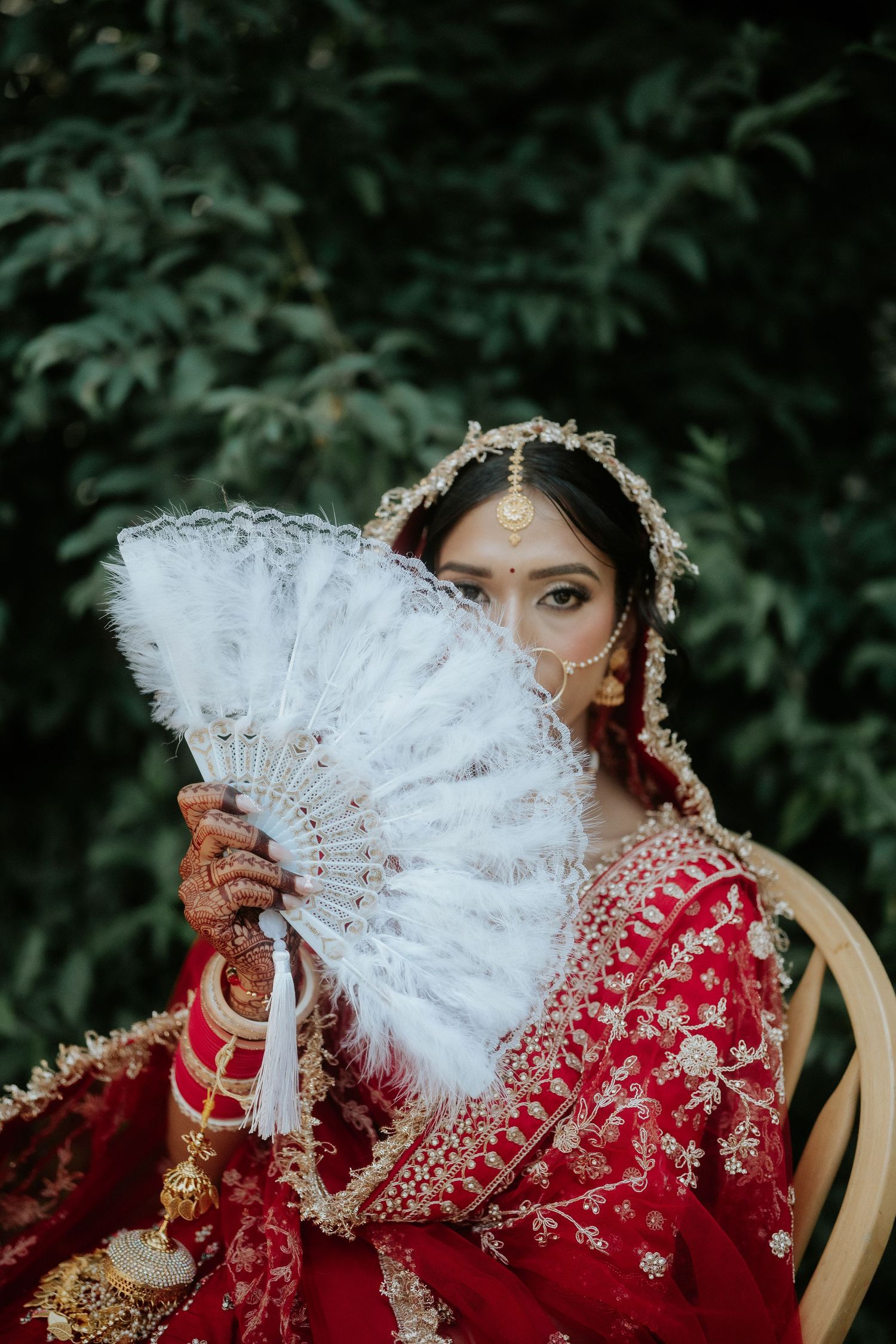 A bride in traditional red Indian attire playfully poses with a decorative white fan outdoors.