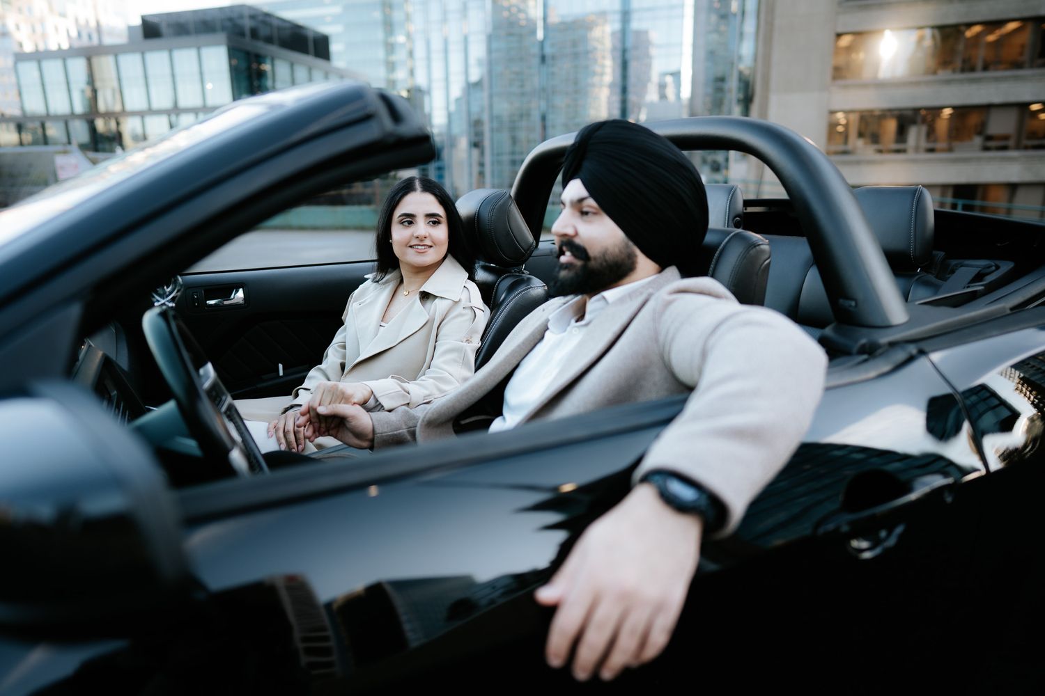 Two people sit in a sleek black convertible car parked in an urban setting with modern buildings in the background.