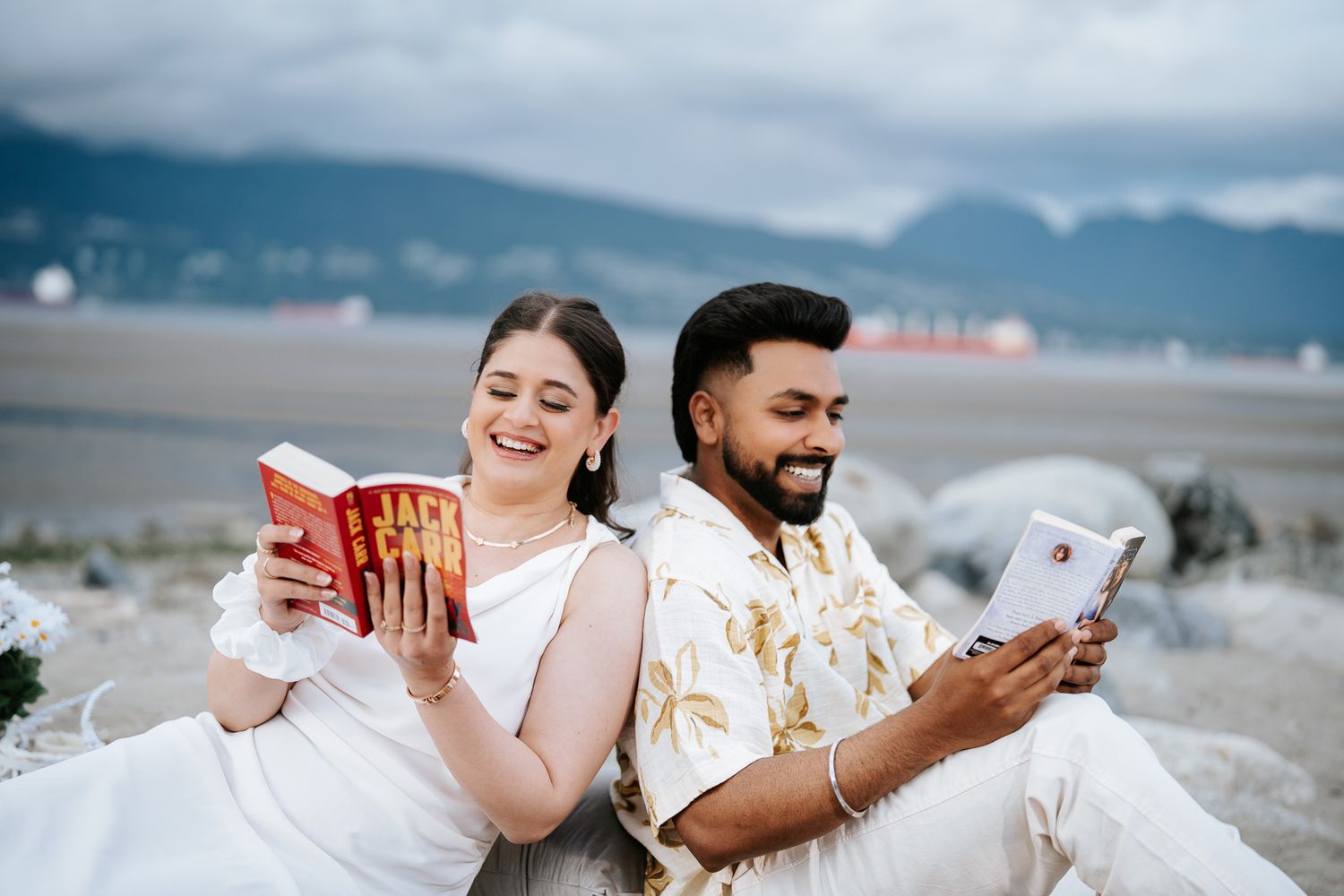 Couple sitting back-to-back on a beach, woman reading a red book and man reading on a tablet, mountains in background.