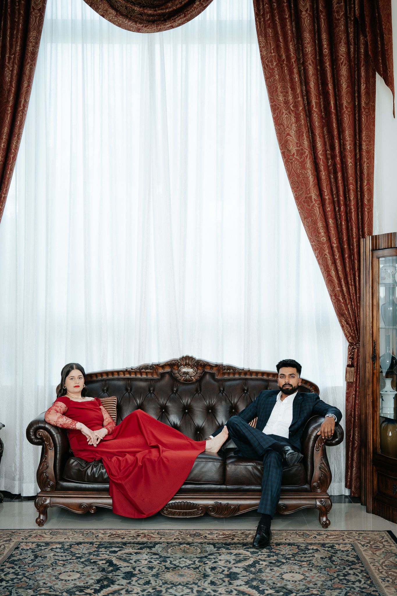 Elegant couple relaxing on tufted leather sofa with burgundy drape curtains backdrop.