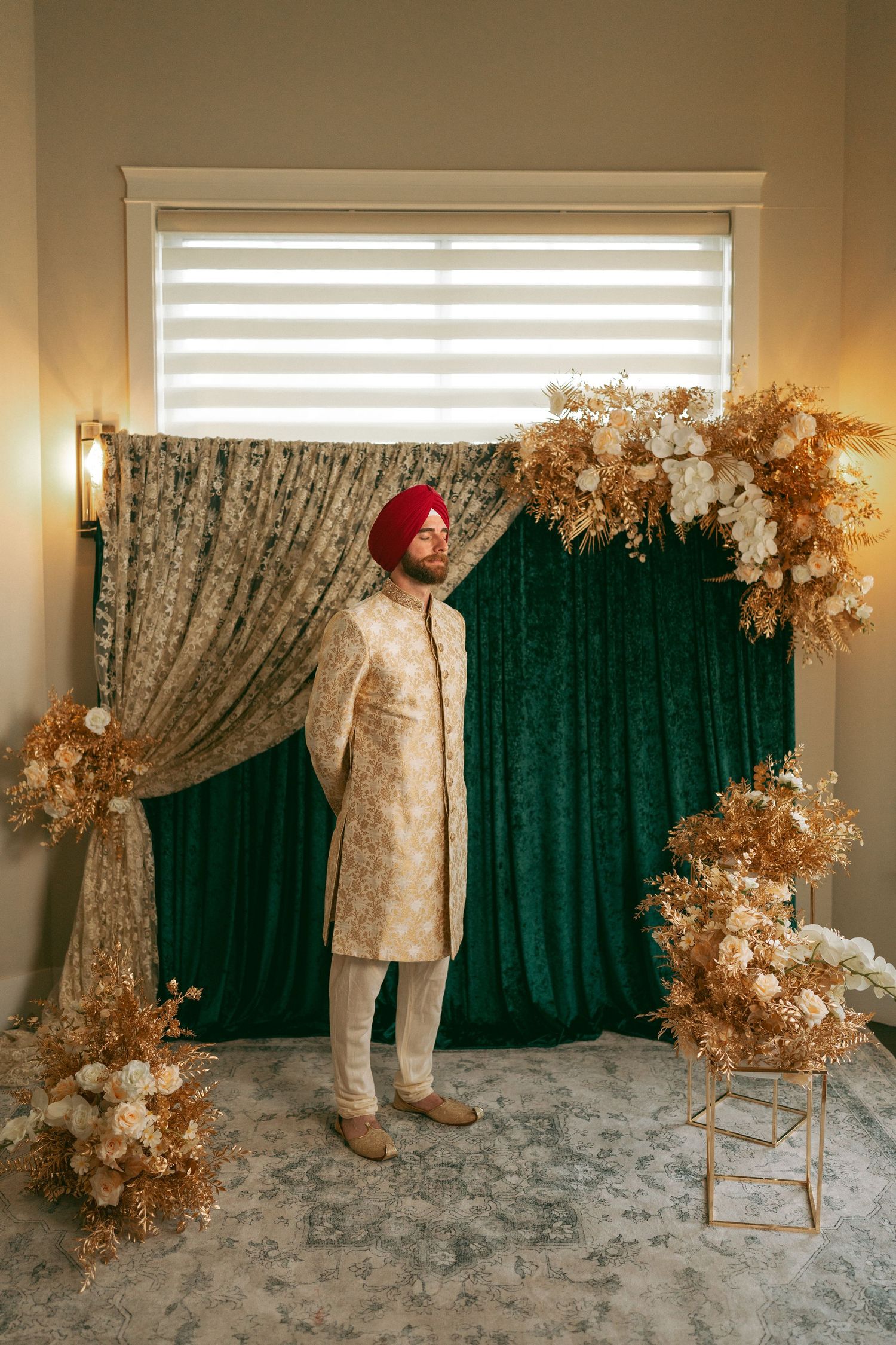 Sikh groom in cream sherwani and red turban poses before green velvet backdrop with gold floral arrangements.