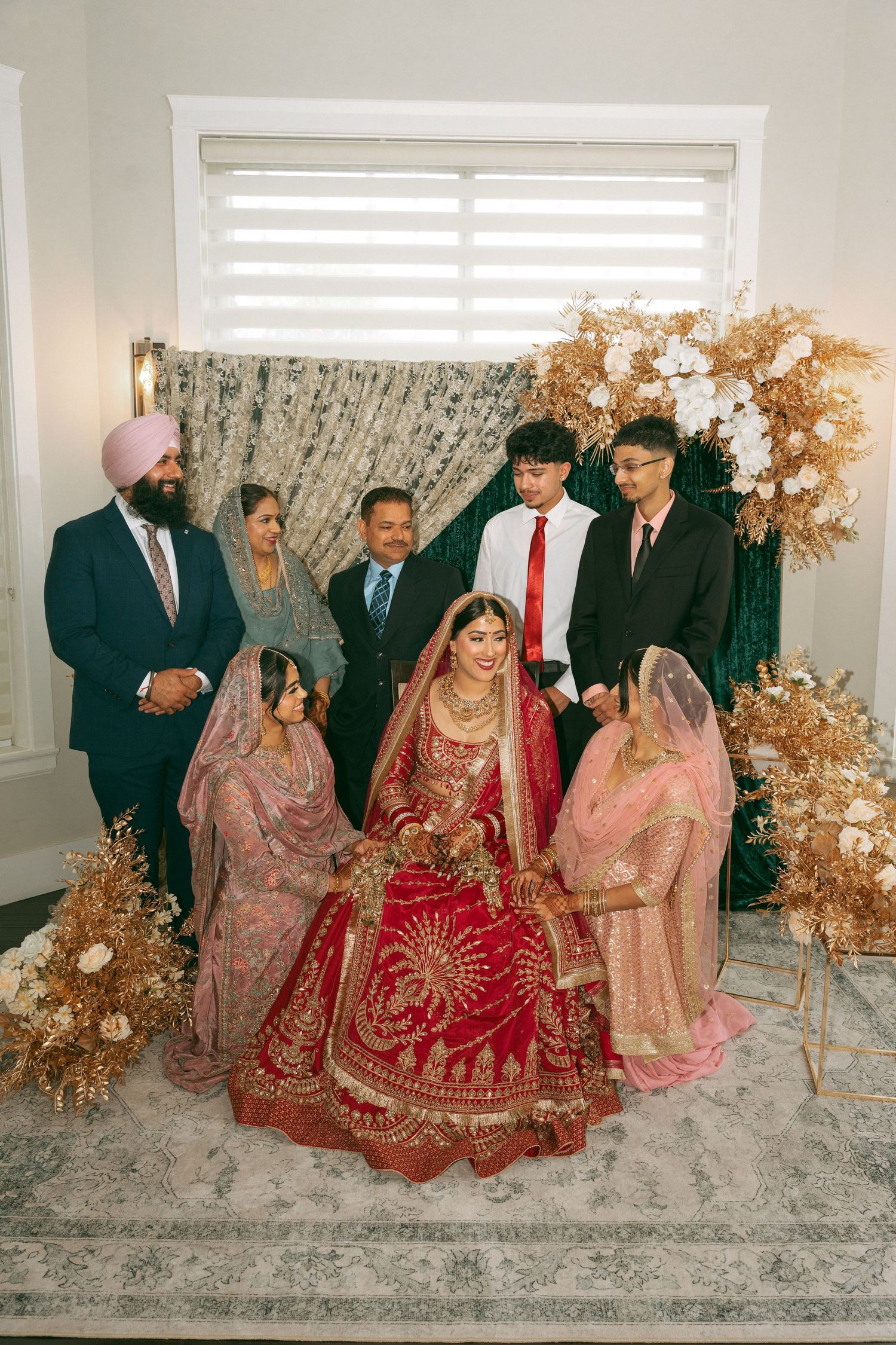 Bride in red lehenga with gold jewelry poses with family at elegant South Asian wedding ceremony indoors.