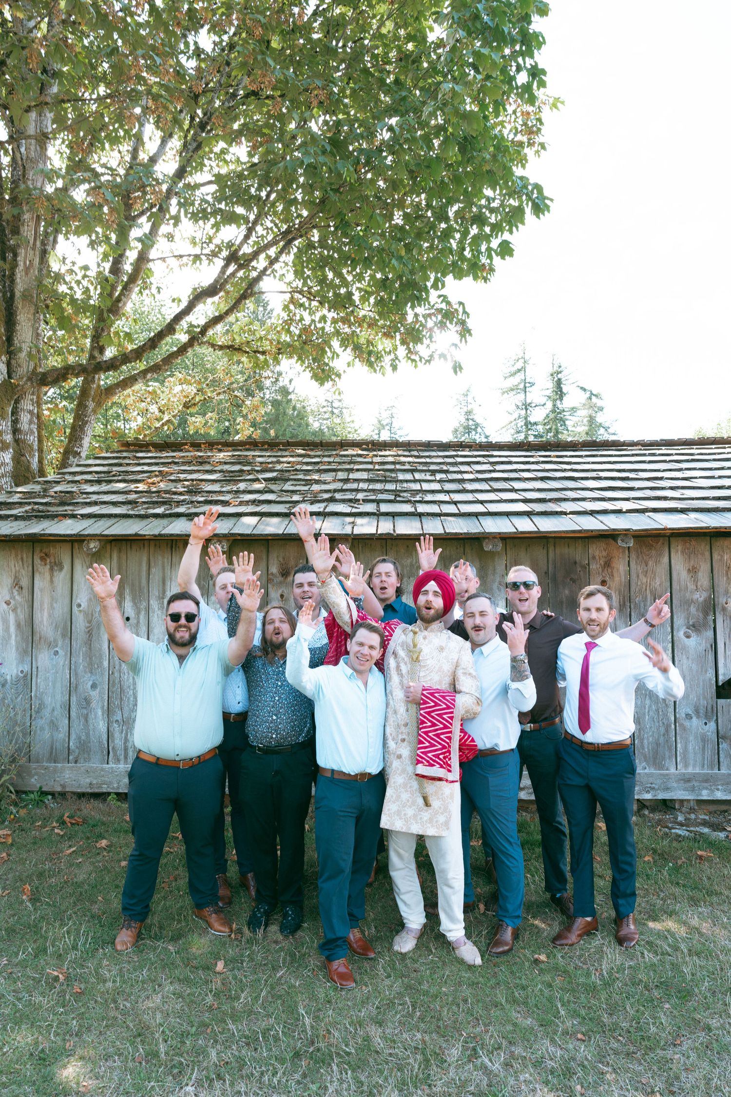 Groom in traditional Sikh attire poses with groomsmen in front of rustic wooden barn, all raising hands joyfully.