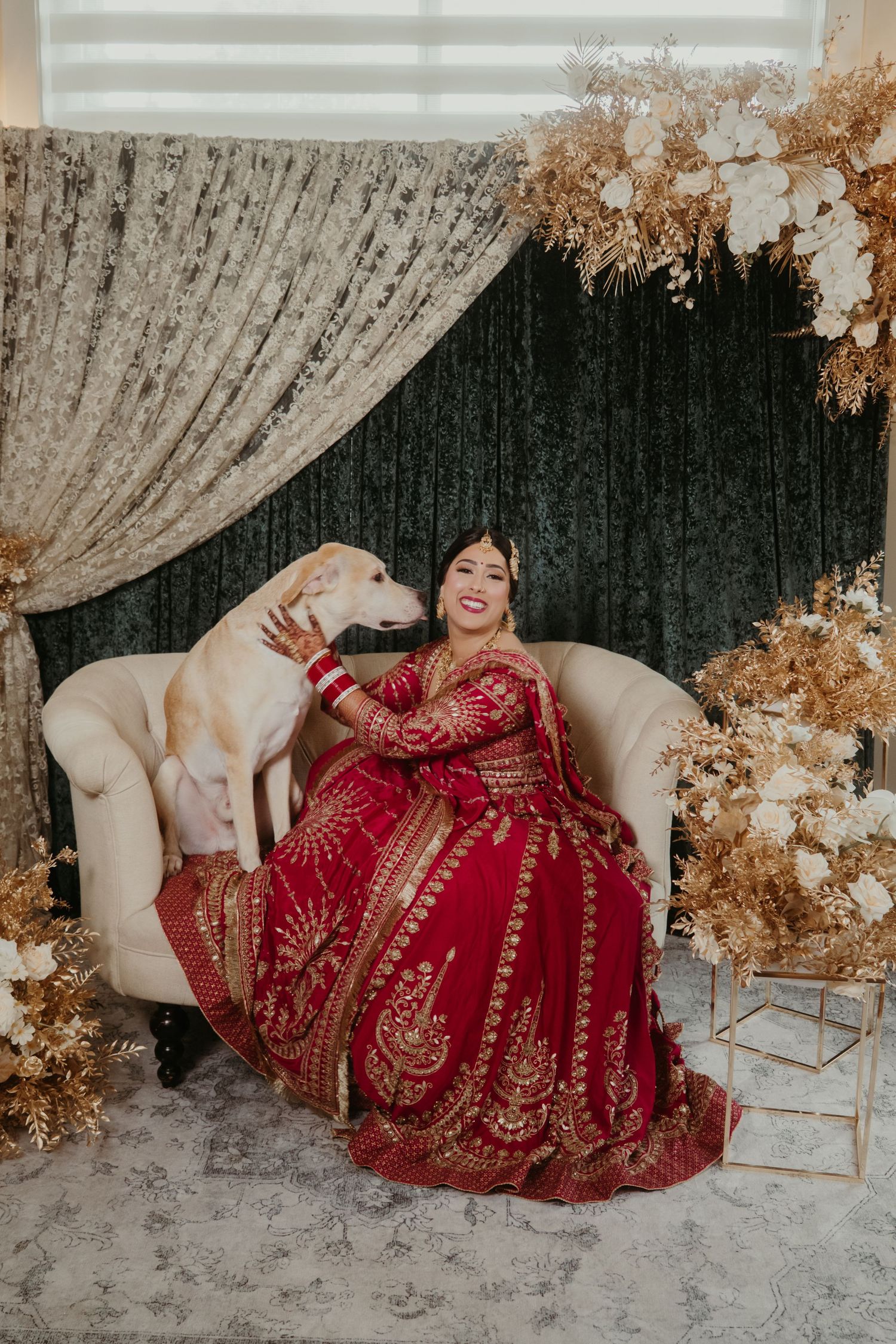 Bride in red and gold embroidered lehenga sits on cream sofa with white dog, surrounded by golden floral decor.
