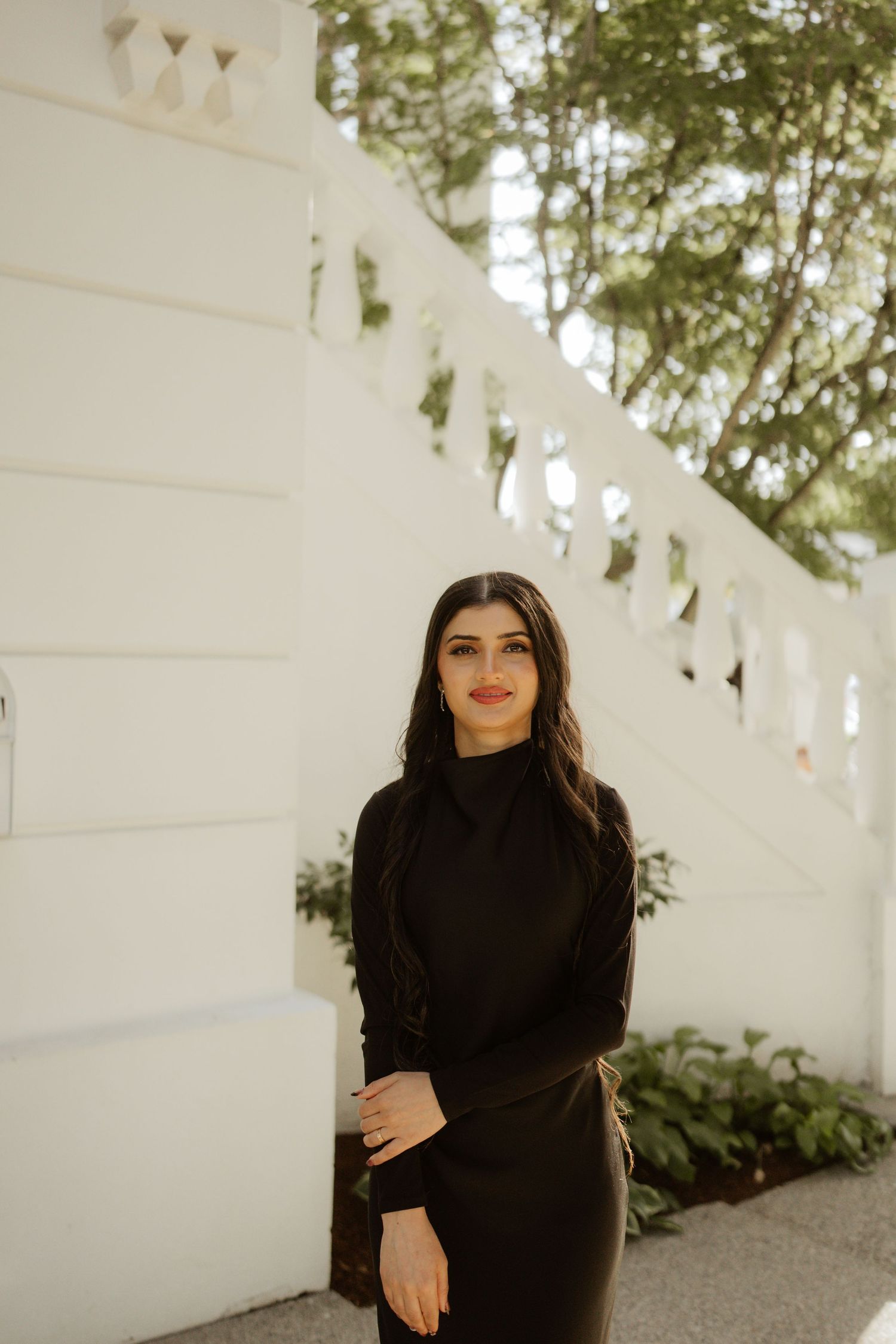 Young woman in black turtleneck and skirt poses near white balustrade wall with green trees background.