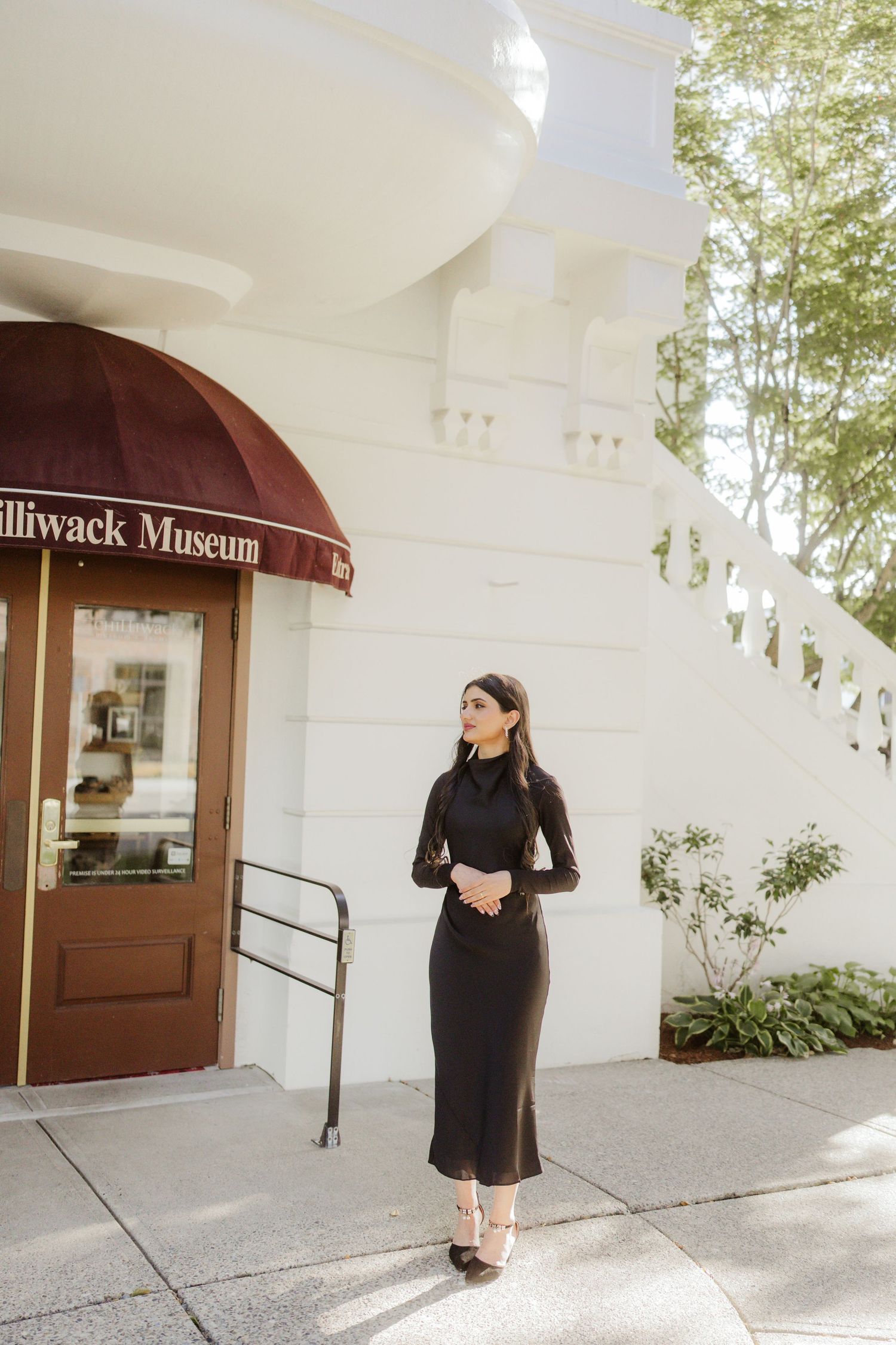 Elegant woman in black midi dress standing outside the Chilliwack Museum on a sunny day.