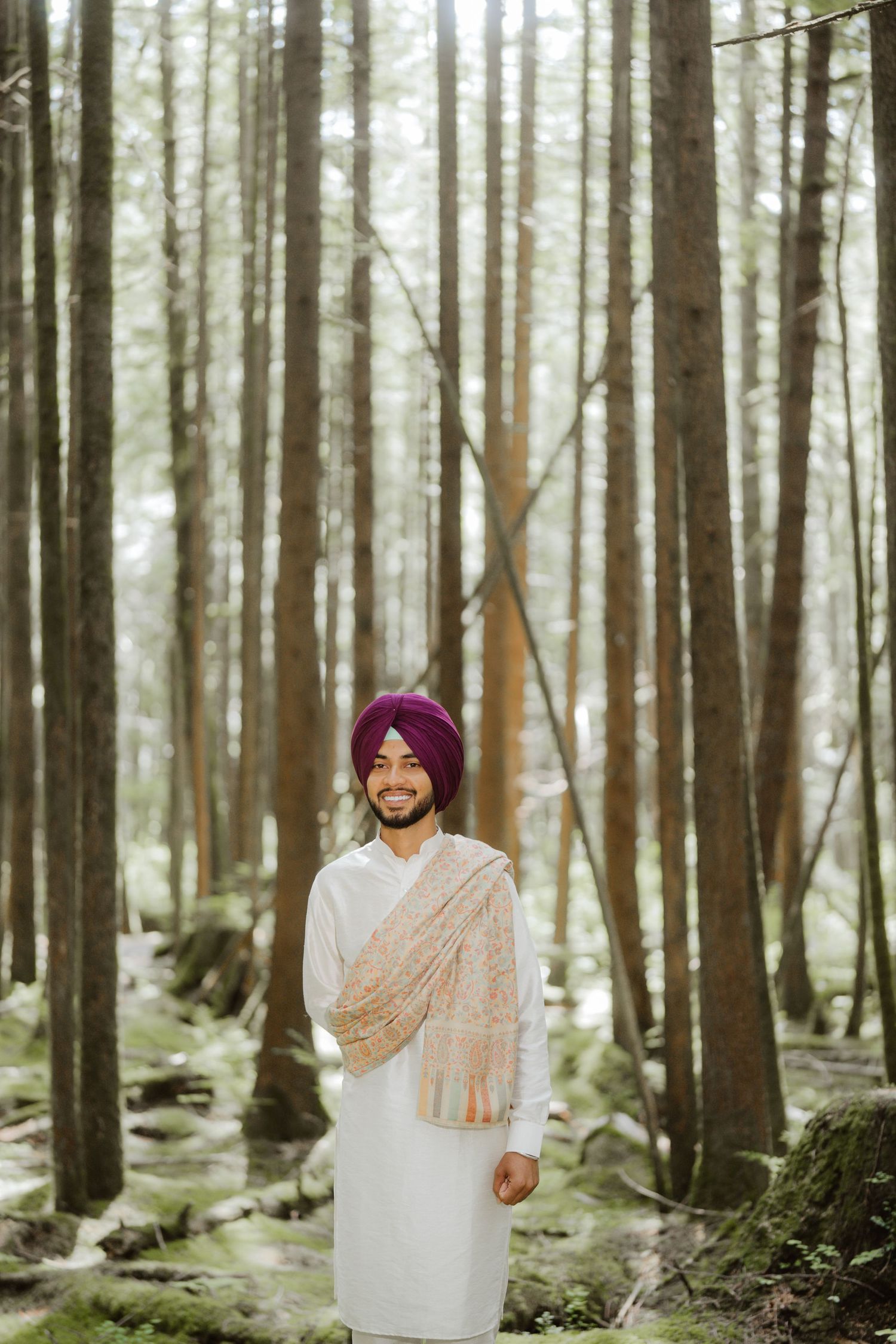 Smiling Sikh man in white kurta and purple turban standing among tall trees in a lush green forest.