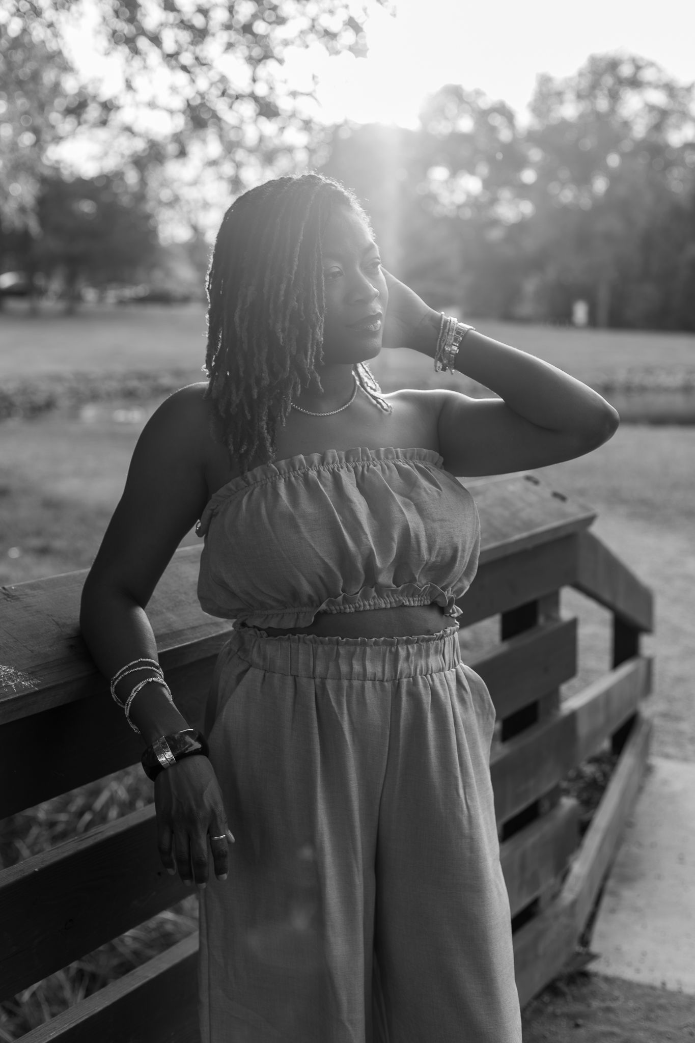 Black and white portrait of a woman in a strapless jumpsuit leaning on a wooden fence in a sunlit park.