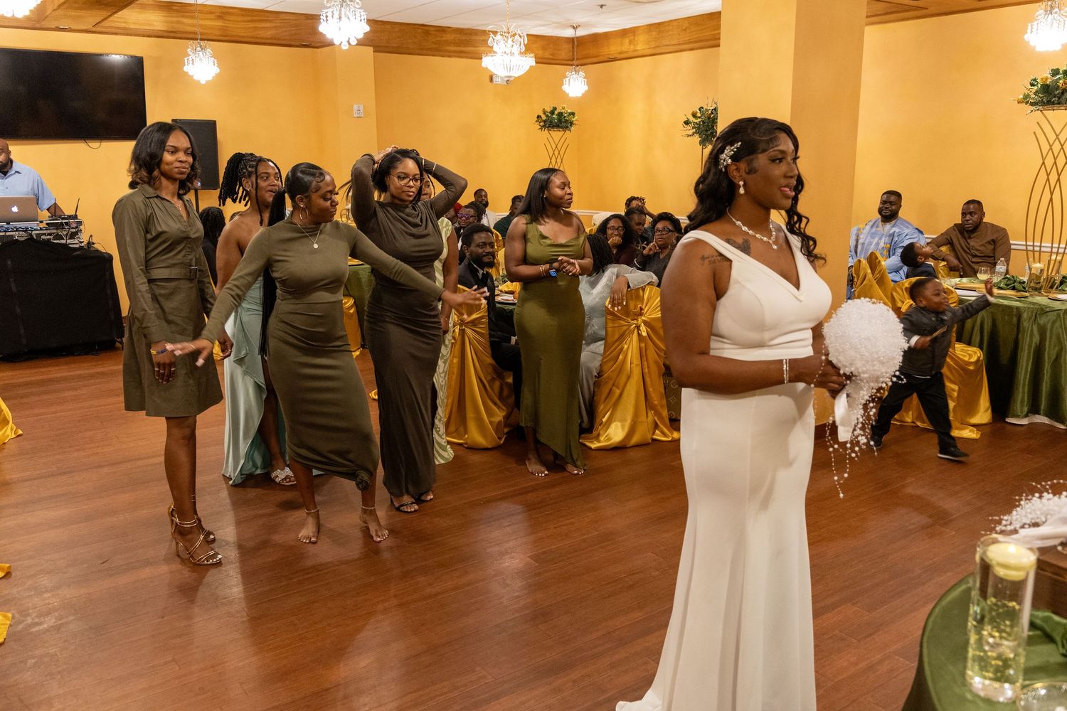 Bride in white gown prepares to toss bouquet to excited bridesmaids in olive dresses at wedding reception.