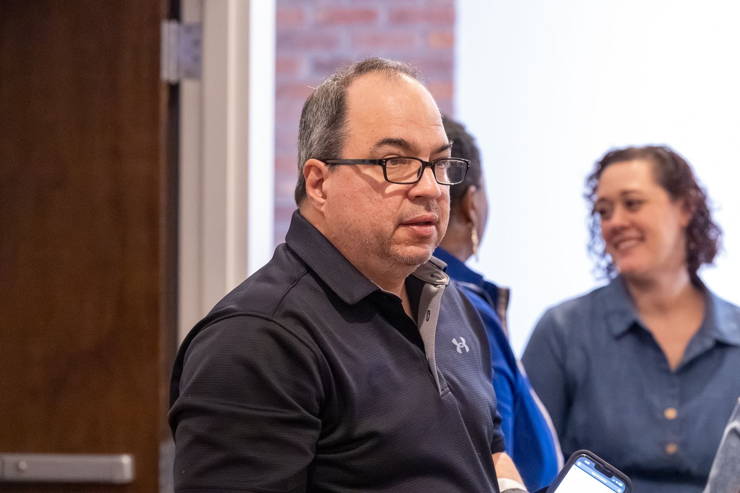 Man in black shirt with glasses speaking at an indoor meeting with colleagues in the background.