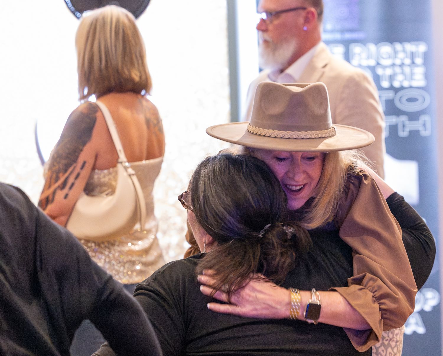 A woman wearing a tan fedora hat warmly embraces another person at an indoor social gathering or event.