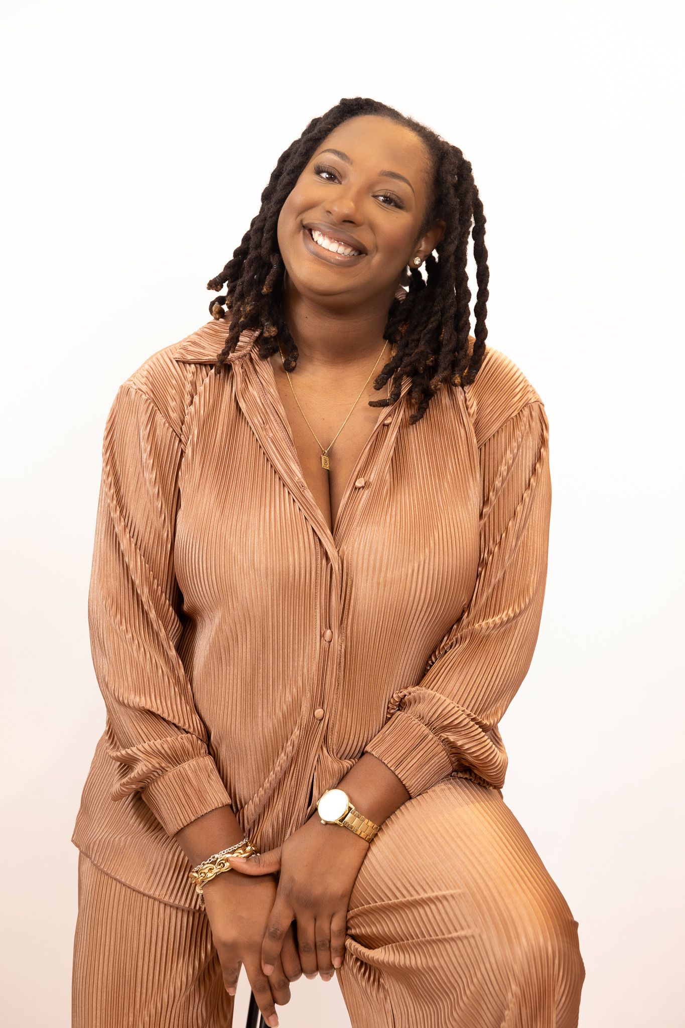 A Black woman with locs wearing a bronze pleated outfit sits smiling broadly with hands clasped in her lap.