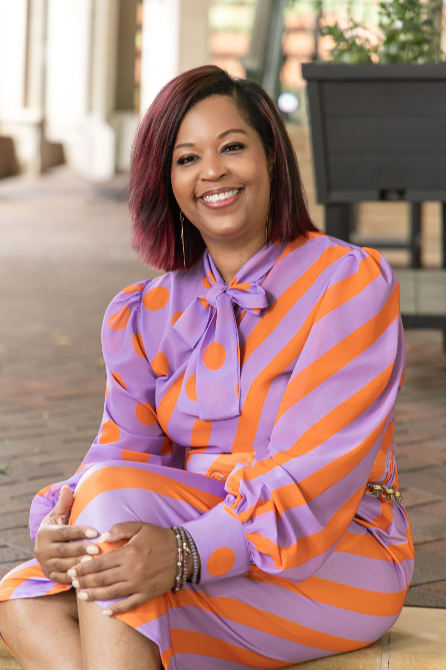 Smiling woman in vibrant purple and orange striped polka dot dress sitting outdoors on brick steps.