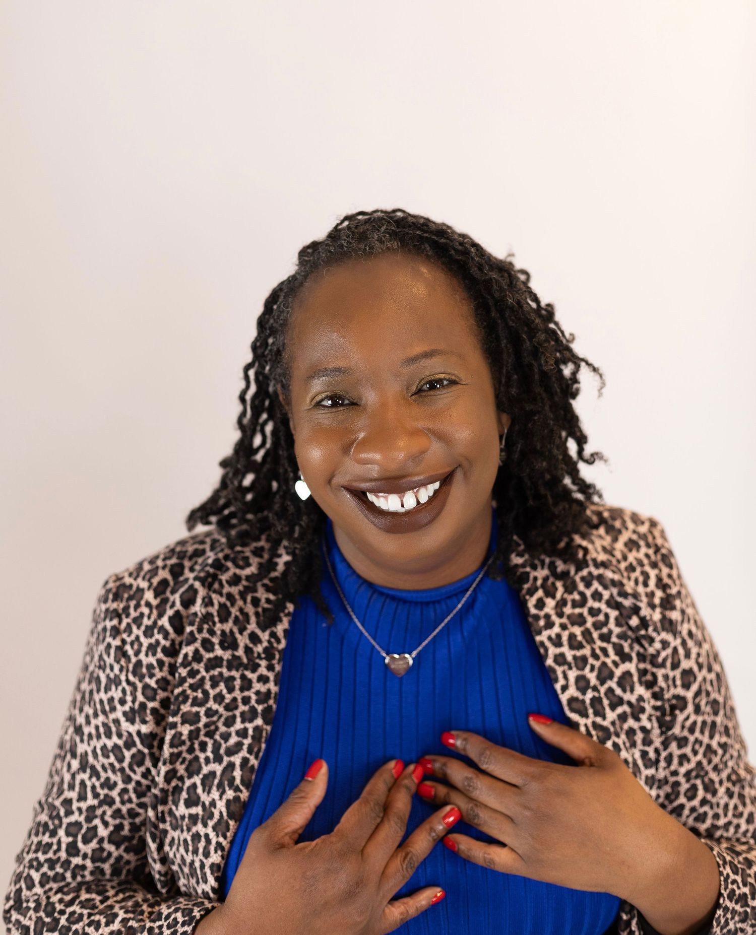 A cheerful Black woman in a blue top and leopard blazer smiles warmly with hands on her chest.