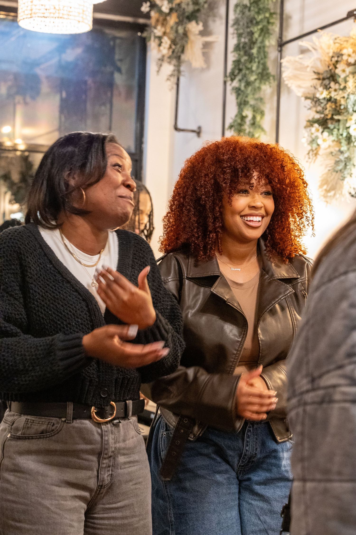 Two women smiling and laughing together at an indoor social gathering with floral decor.