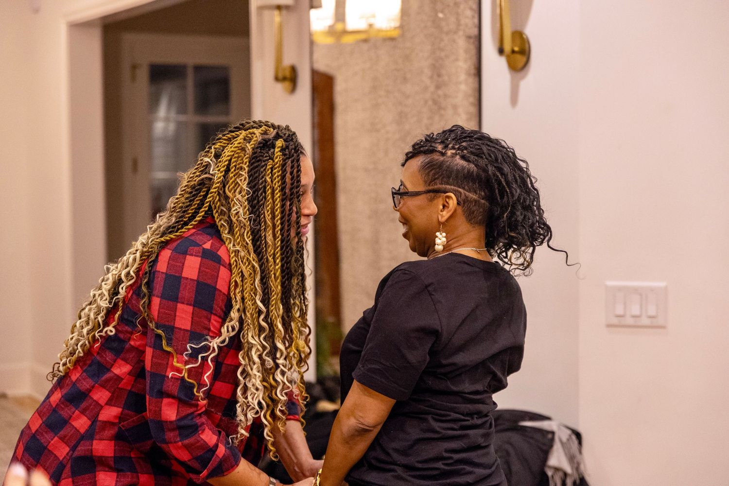 Two women with colorful braided hairstyles share an intimate conversation at an indoor gathering.