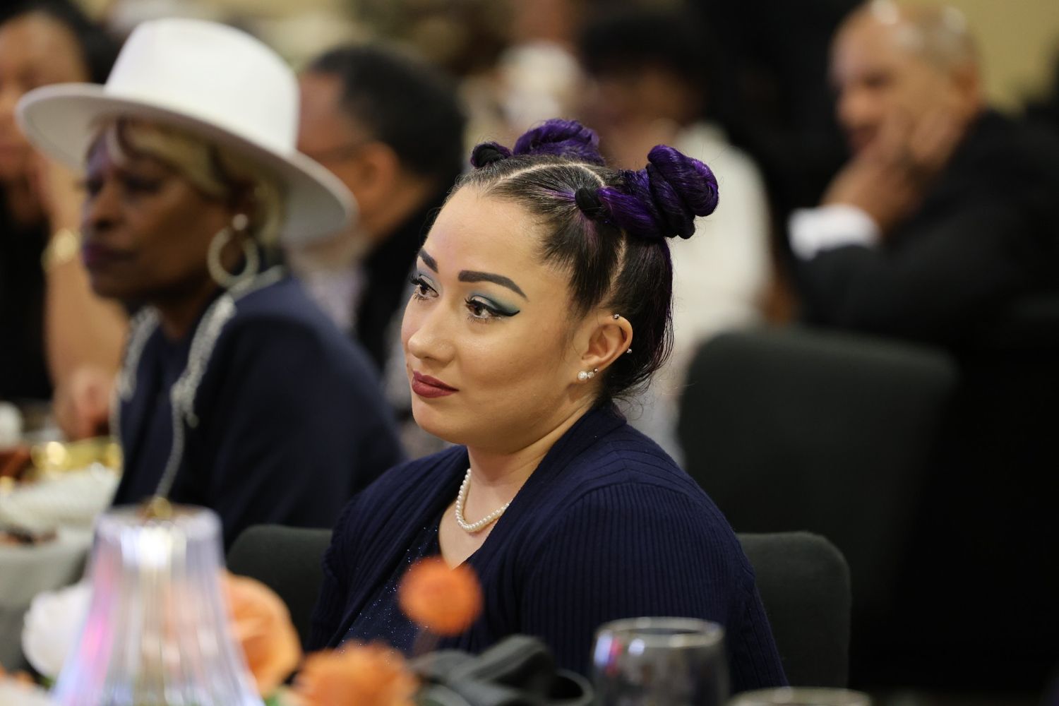 A woman with purple hair buns and dramatic makeup sits at a formal banquet table.