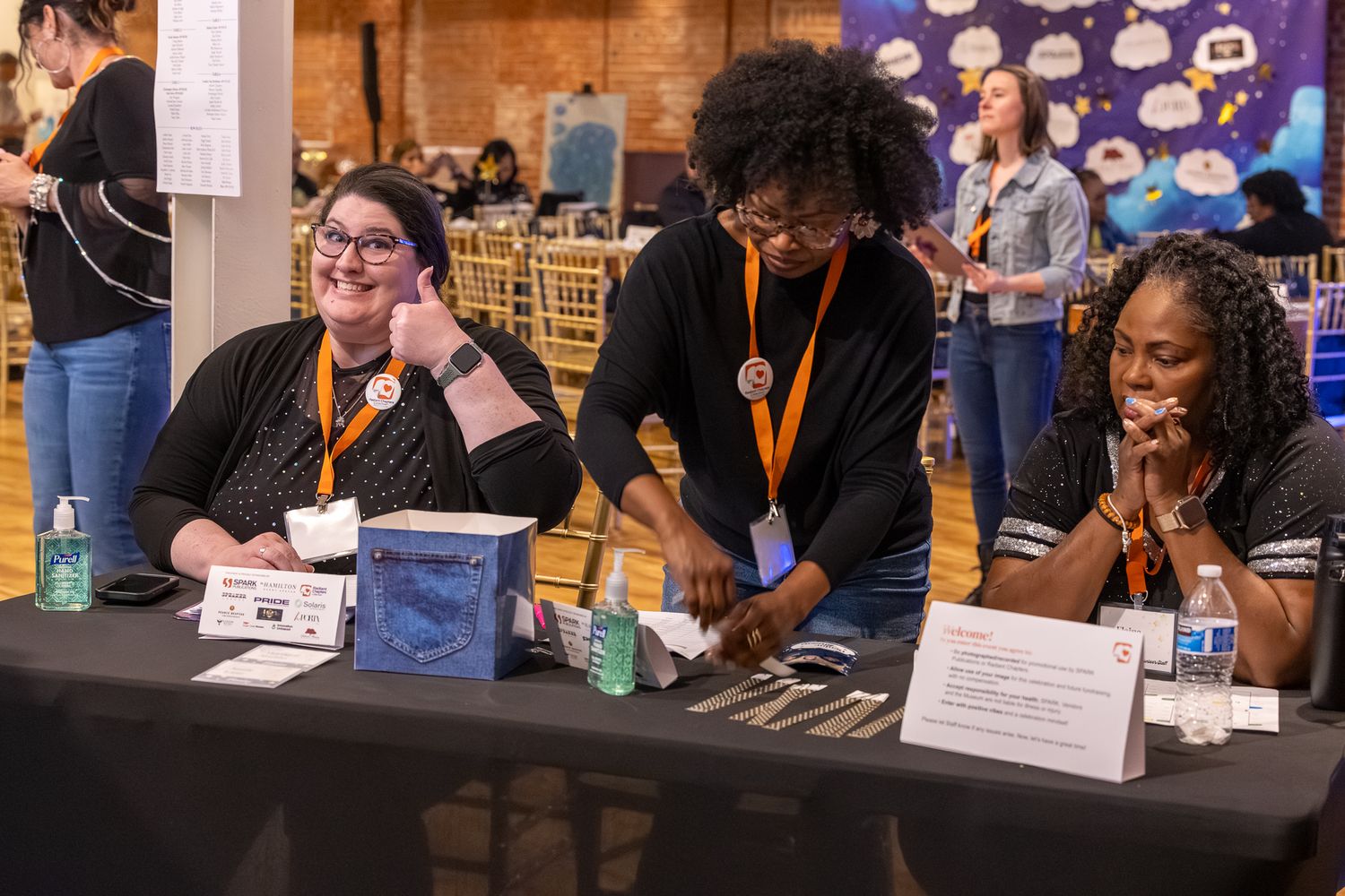 Three women wearing orange lanyards smile and work together at a table during a professional conference or event.