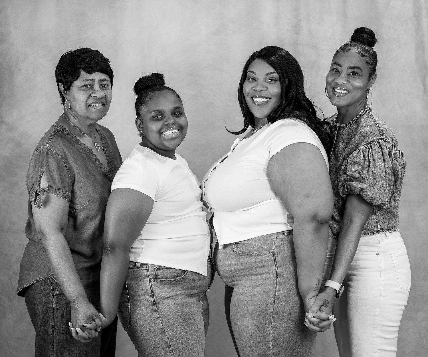 Four women smile and hold hands in a black and white portrait photo studio session.