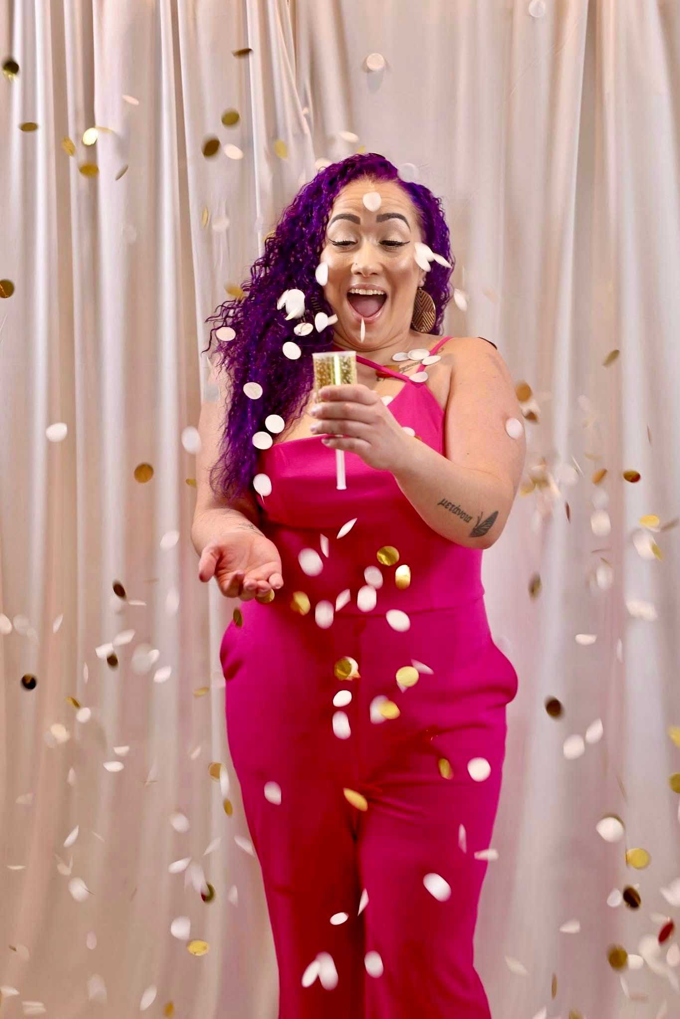 Joyful woman in pink jumpsuit celebrating with confetti and champagne against white curtain backdrop.