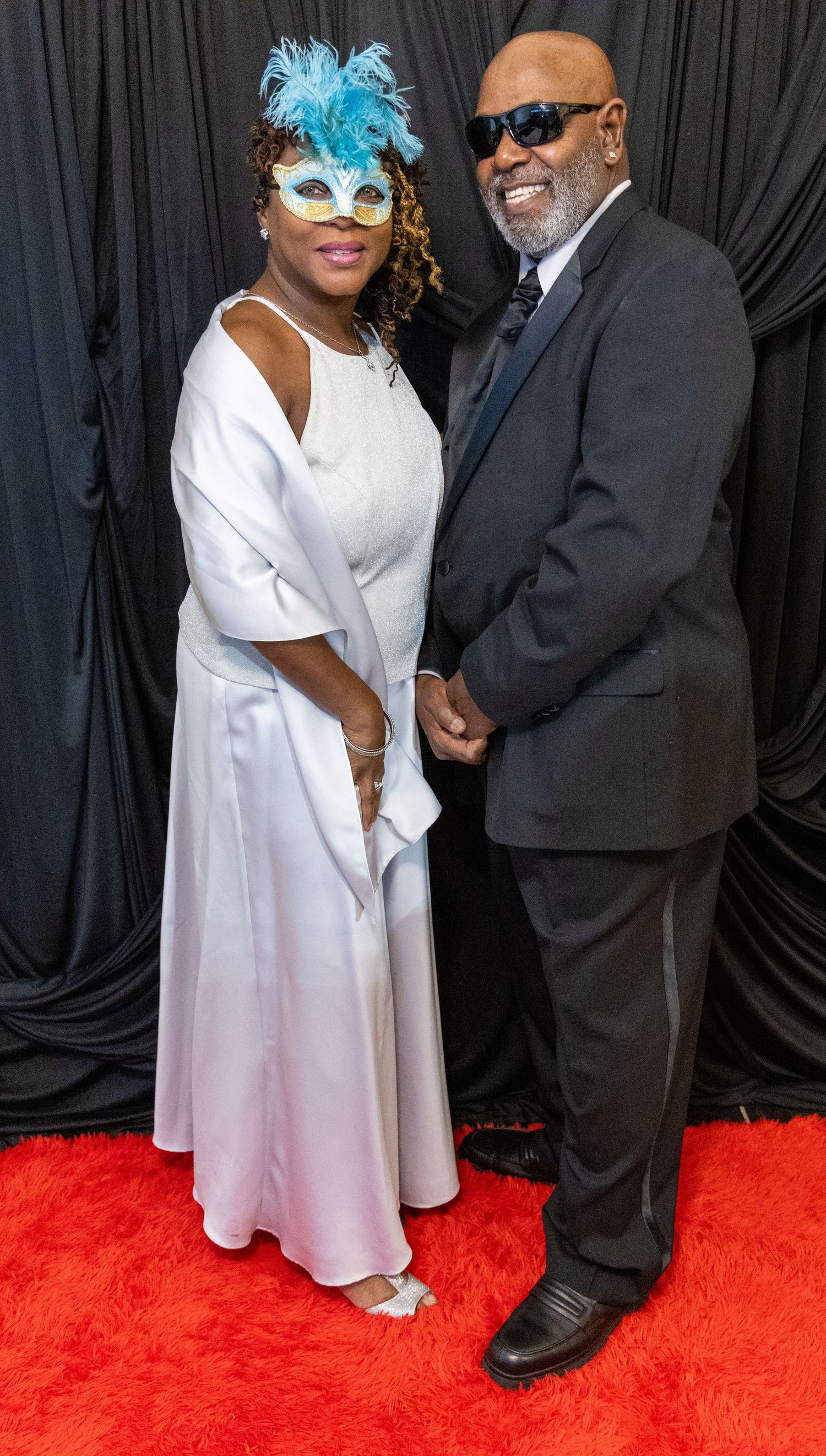 Couple posing on red carpet at masquerade gala, woman in white gown with blue mask, man in black suit with sunglasses.