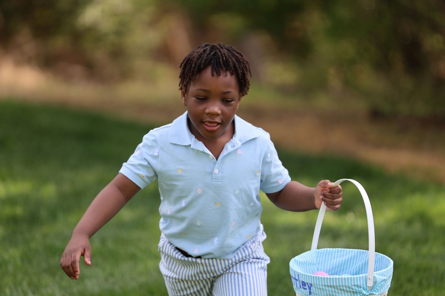 A young Black boy in a white polo shirt carries a blue Easter basket while hunting for eggs in a green park.