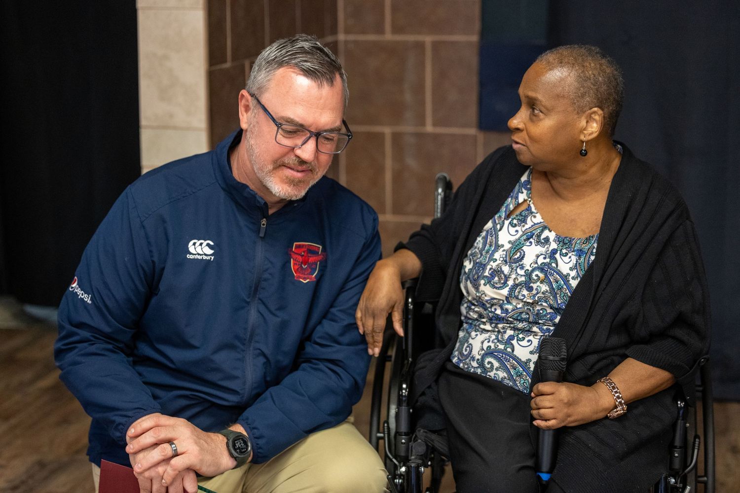 A man in a blue Canterbury jacket converses with a woman in a wheelchair at an indoor event.
