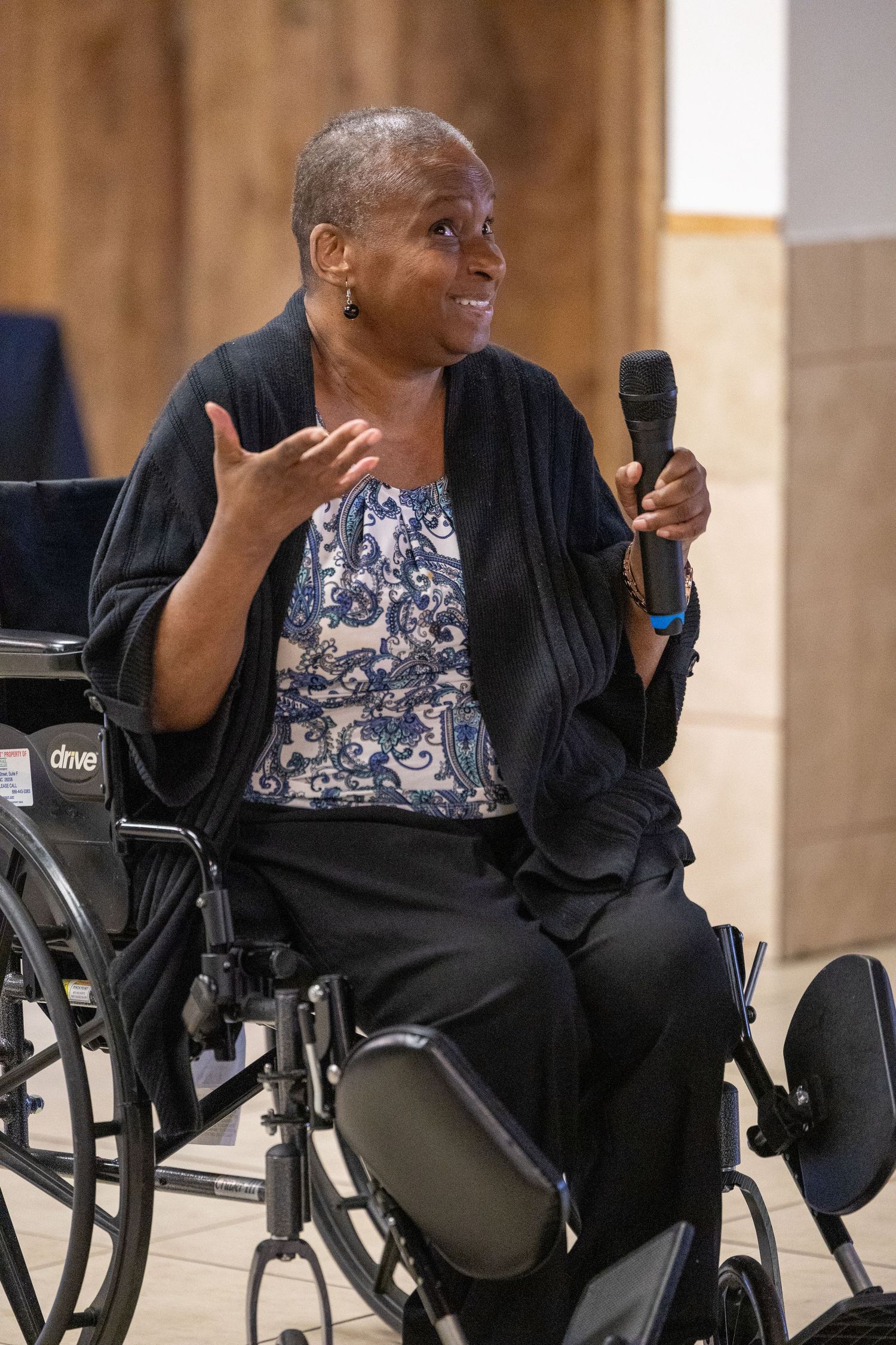 A woman in a wheelchair holds a microphone and gestures while speaking at an event, wearing a black cardigan.