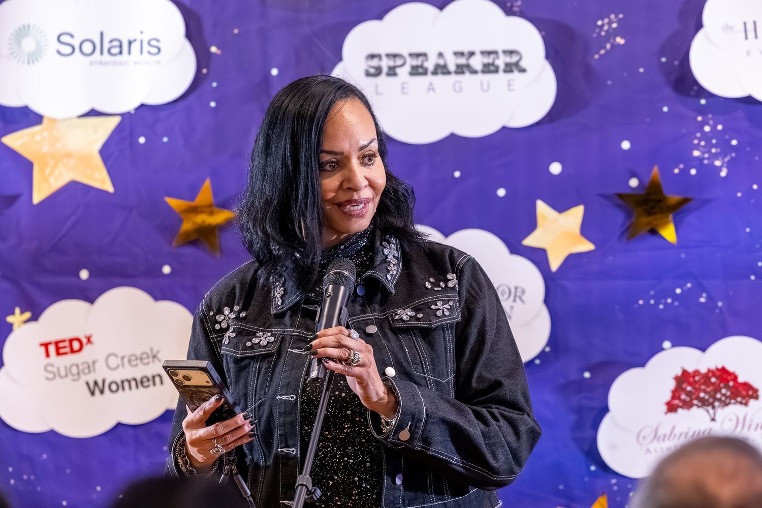 Woman speaking at TEDx Sugar Creek Women event, holding phone at star-decorated speaker backdrop.