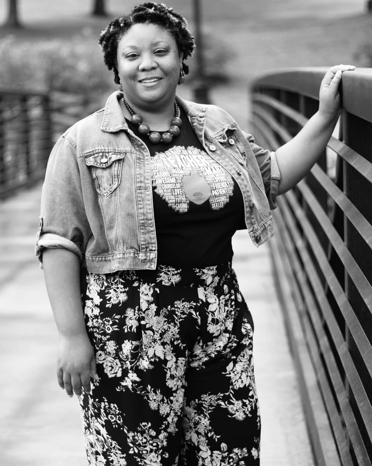 Black and white portrait of smiling woman in denim jacket and floral pants leaning on bridge railing.