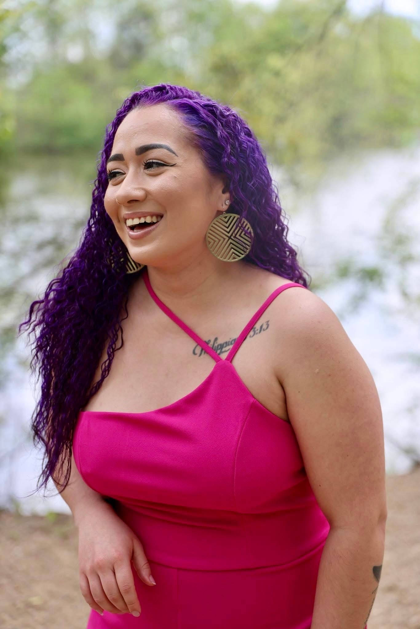 Woman with purple hair in pink dress laughing outdoors near water wearing gold circle earrings.