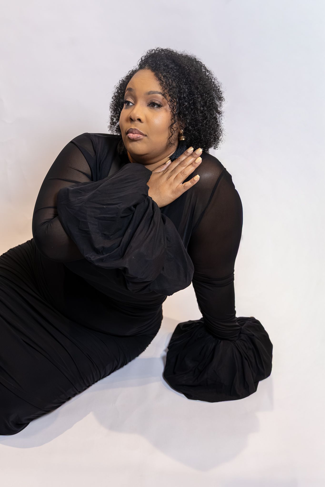 Woman in black sheer dress with dramatic sleeves poses gracefully on white studio floor.