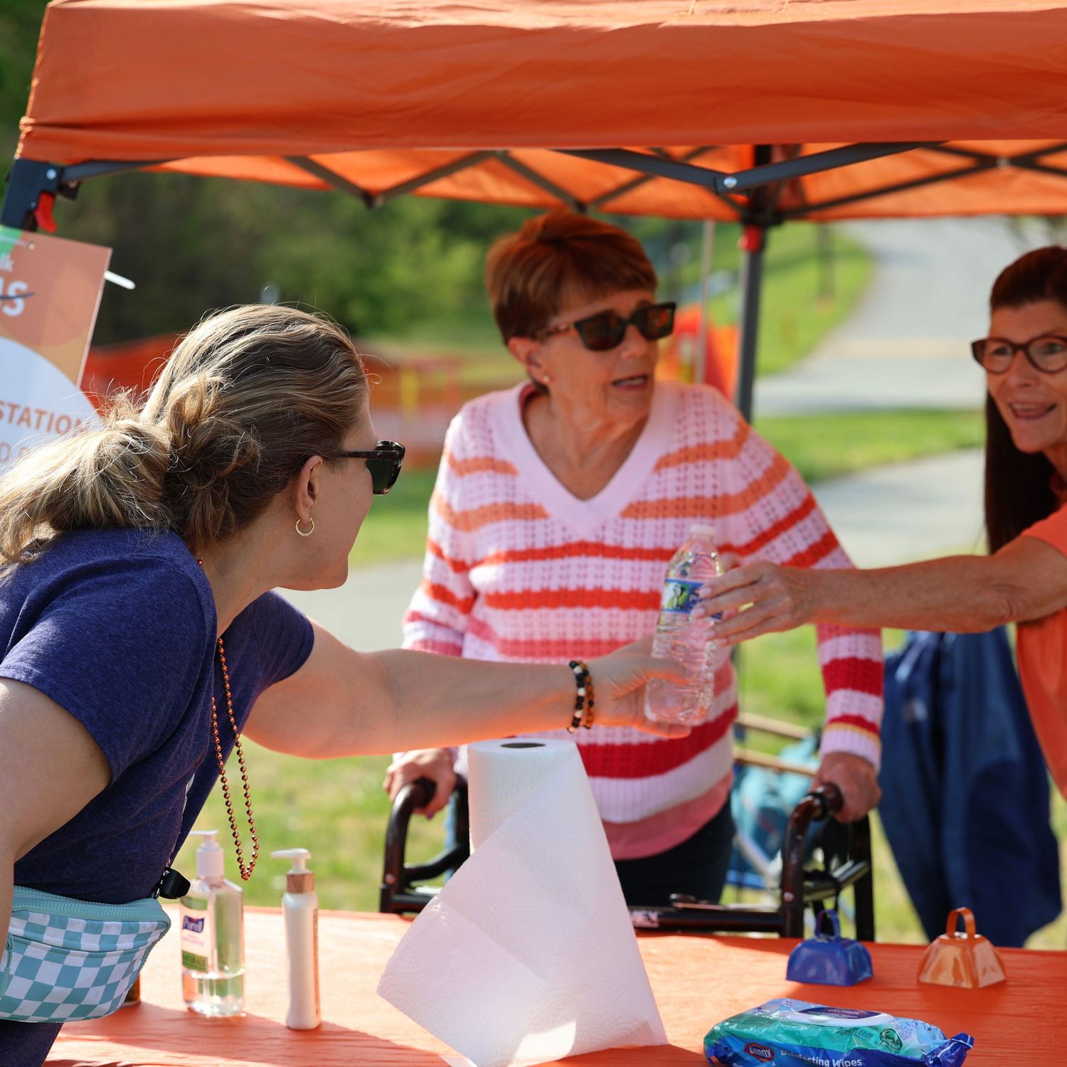 Three women interact at an outdoor event booth under an orange canopy on a sunny day. Multiple Sclerosis. Walk MS
