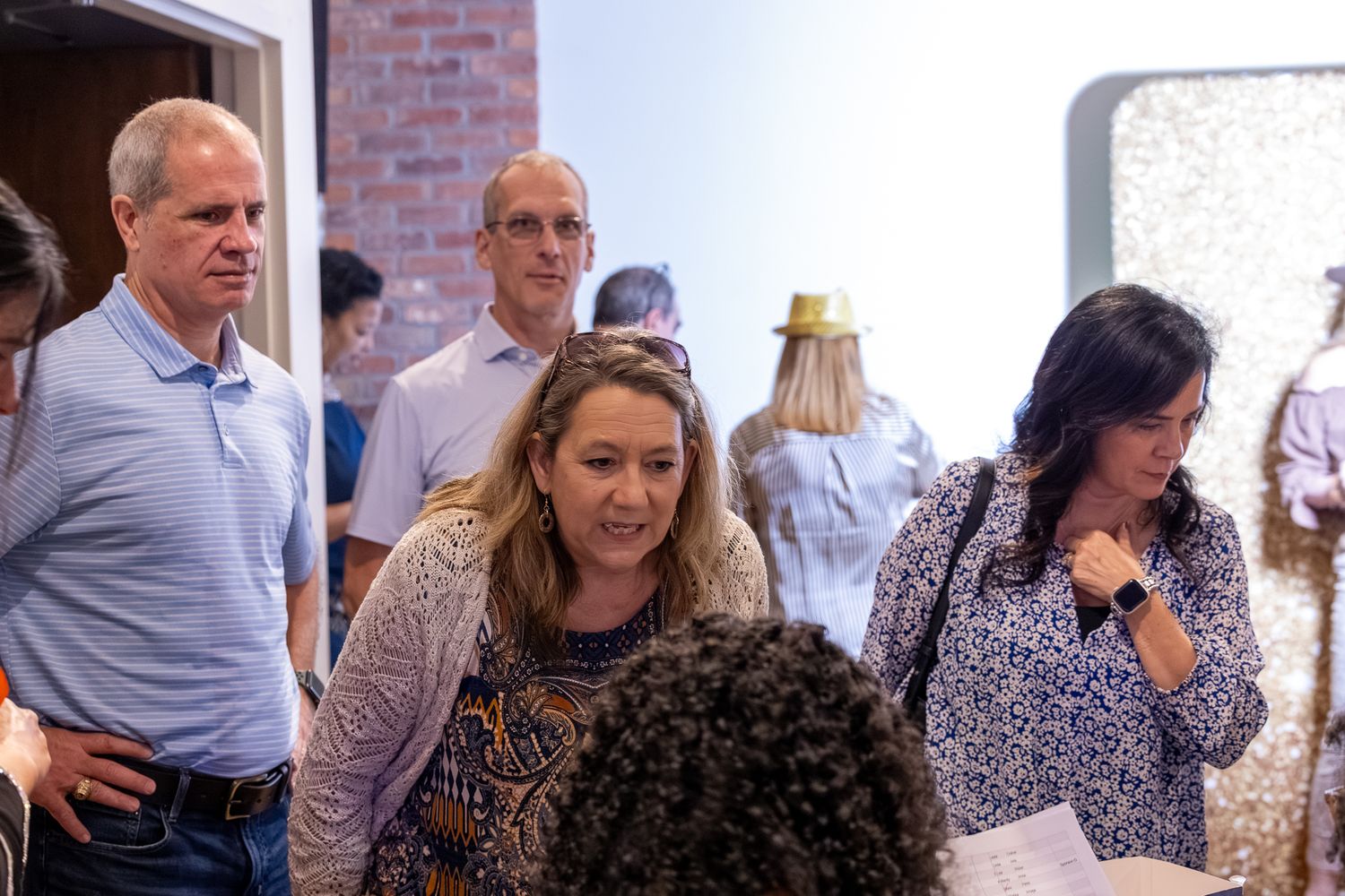 Group of adults mingling at an indoor event, standing near a brick wall in a casual social gathering.