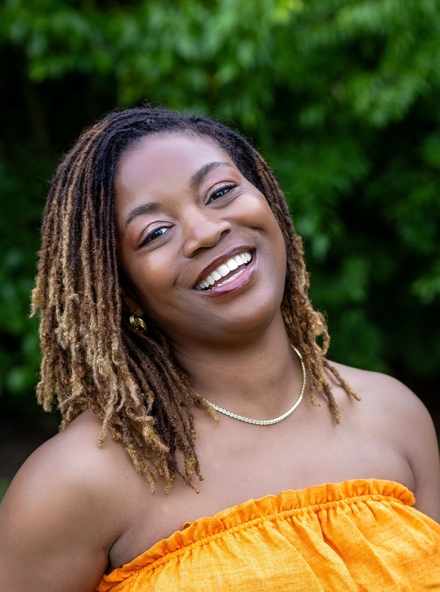 Smiling Black woman with locs wearing an orange off-shoulder top and gold necklace, posing outdoors with green foliage.