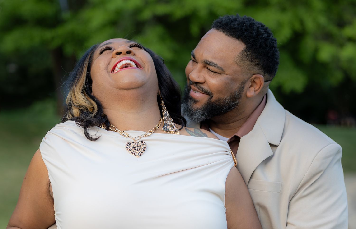 A joyful Black couple shares a loving moment outdoors, woman laughing in white dress with heart necklace, man in beige suit.