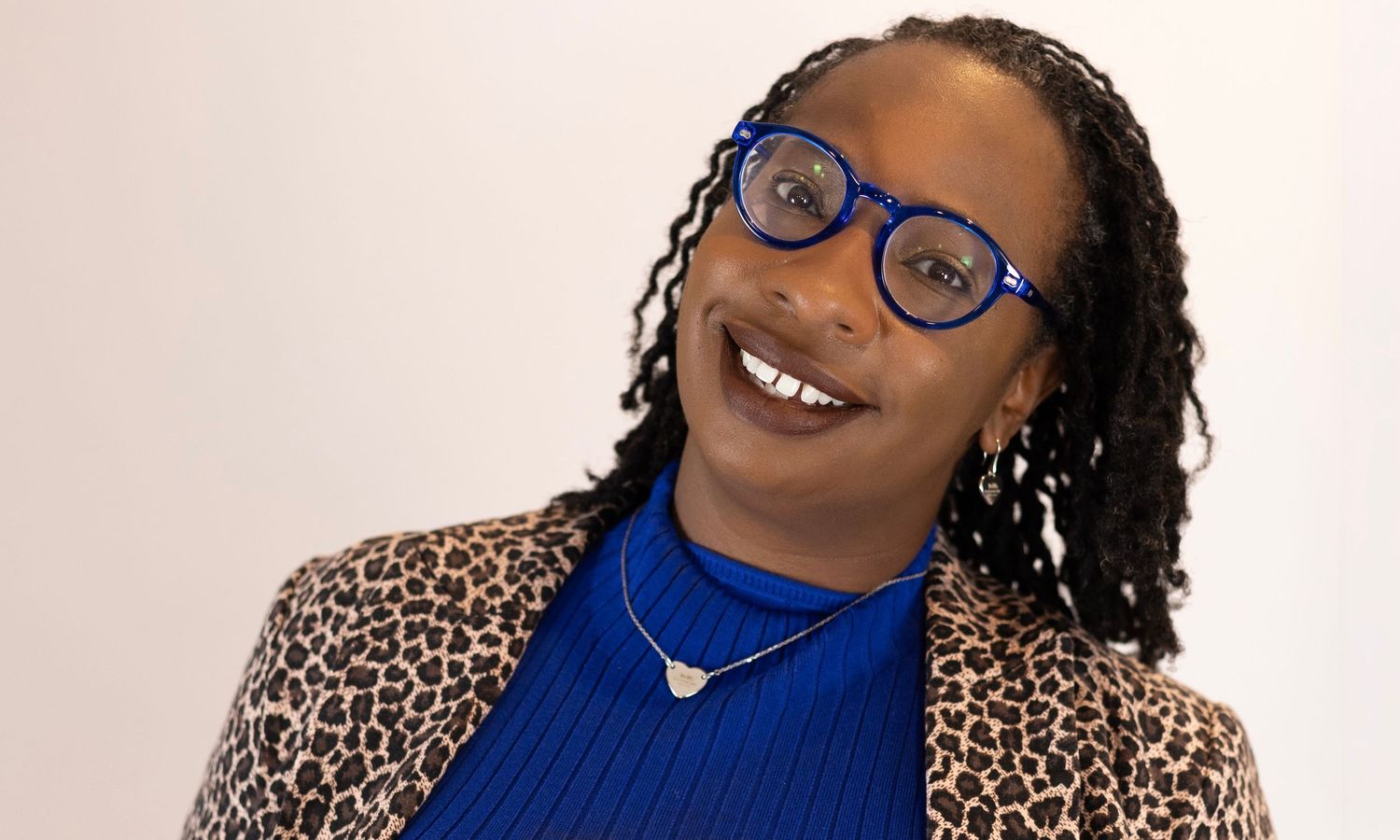 A smiling woman wearing blue glasses and a leopard print blazer over a blue turtleneck poses confidently.