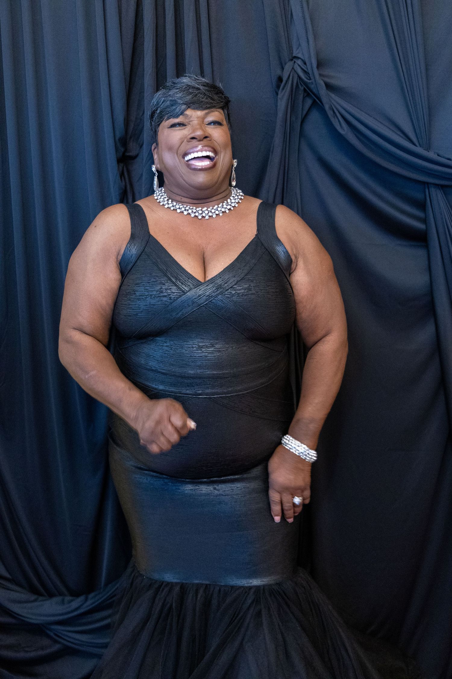 Joyful woman in black leather dress laughing in front of black draped backdrop at event.