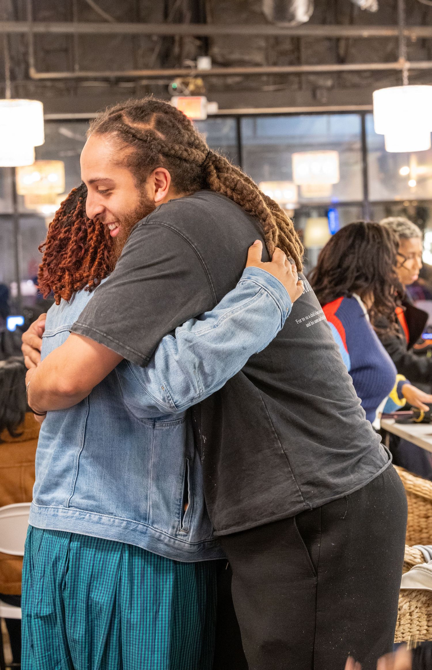 Two people with dreadlocks share a warm embrace, smiling in a lively indoor gathering space.