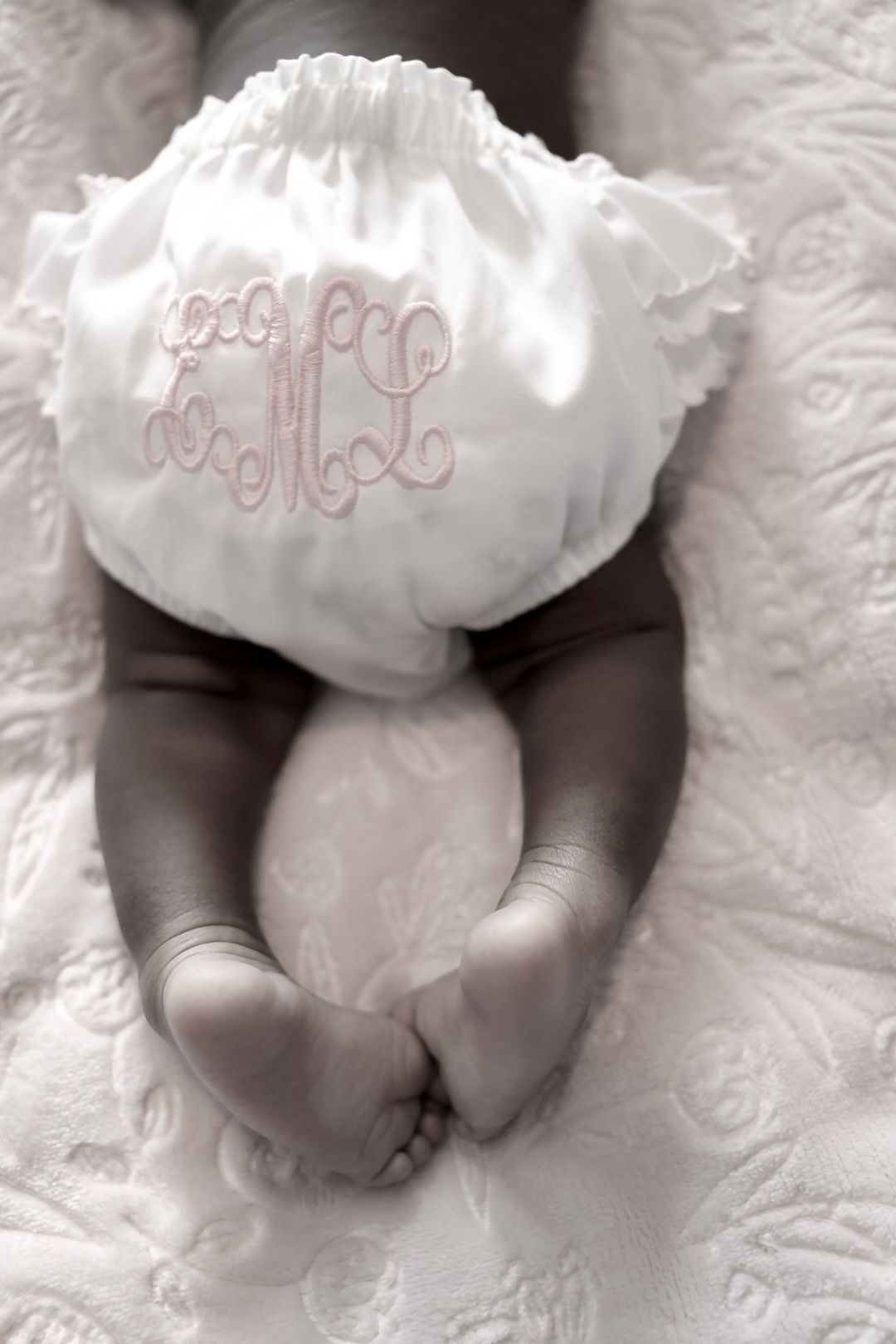 Close-up of newborn baby legs with white monogrammed ruffle diaper cover on textured white blanket.